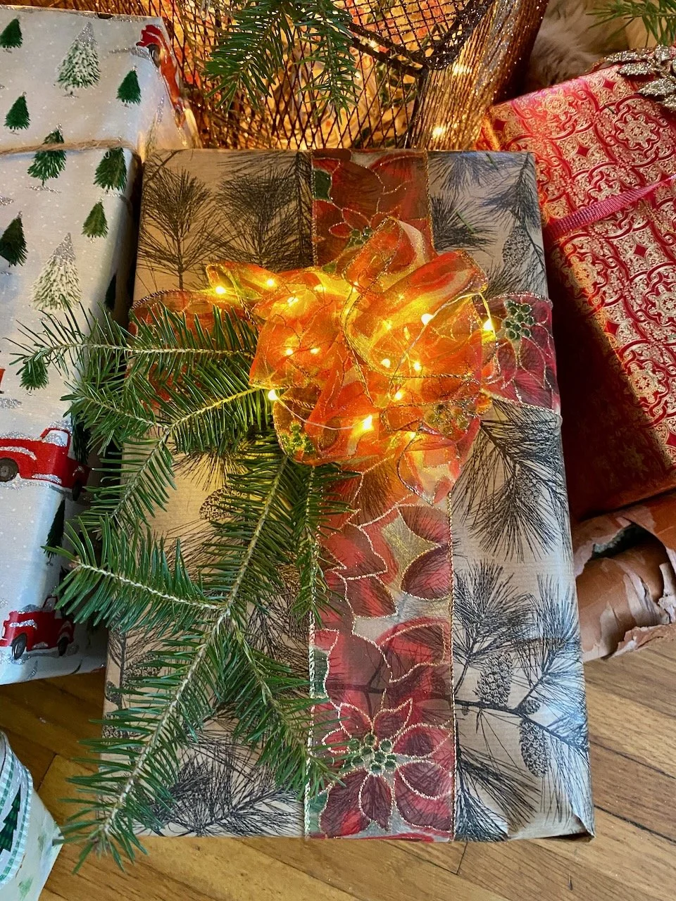 Decorated Christmas gift wrapped in festive paper with poinsettia and pine branch designs, adorned with a glowing amber ribbon and string lights.