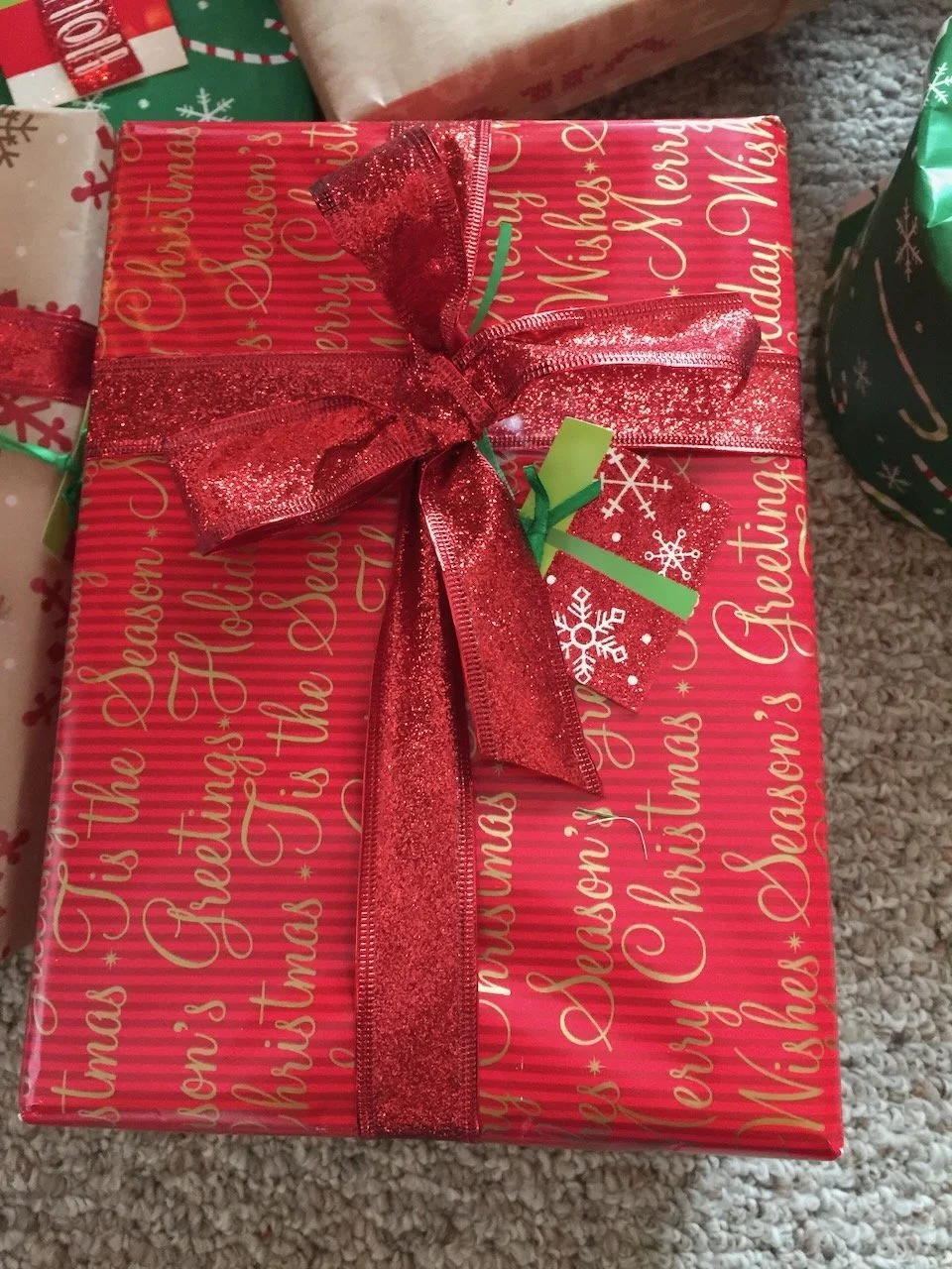 A Christmas gift wrapped in red paper with gold lettering and a red glittery ribbon tied in a bow.