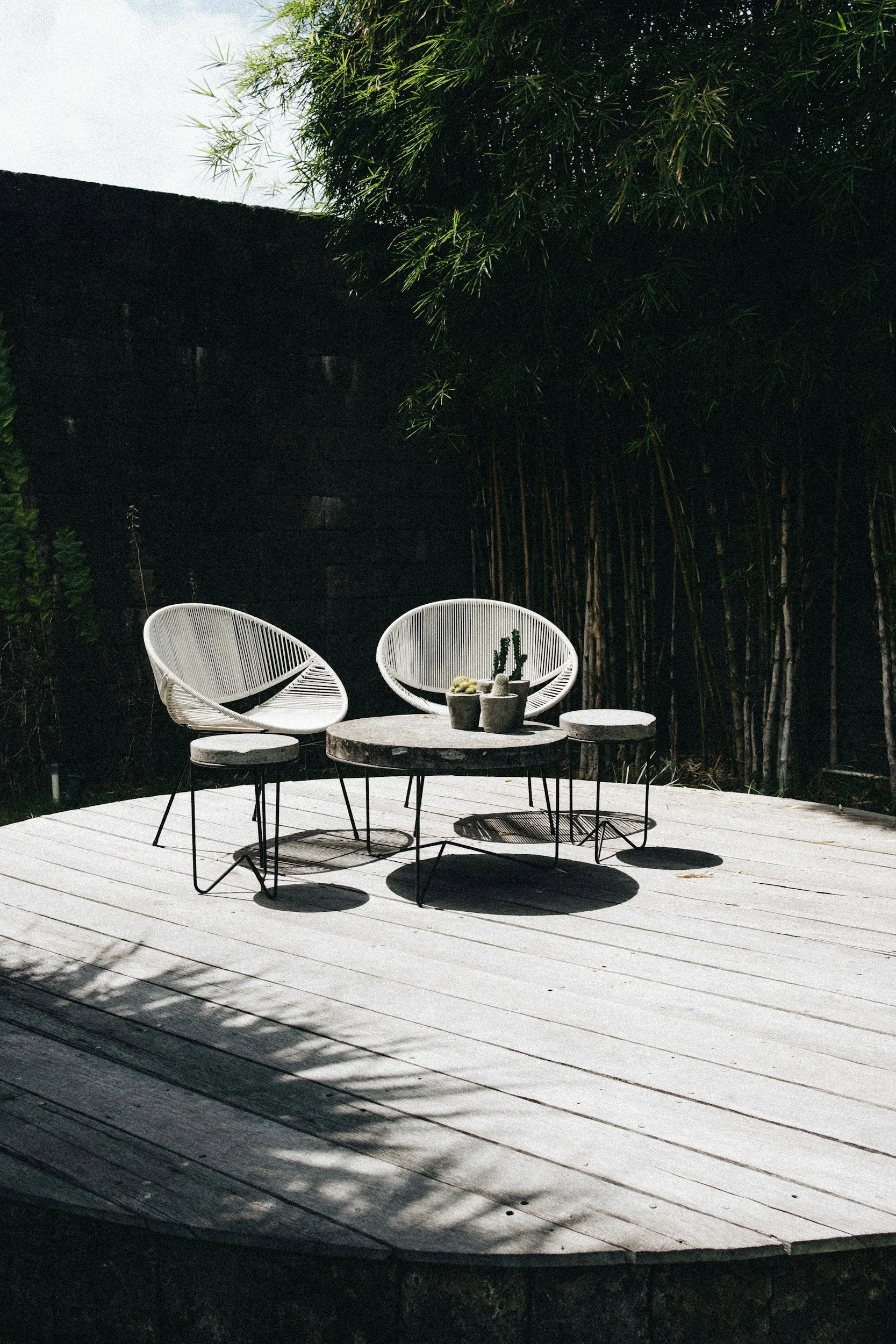 Outdoor patio with round table and chairs, potted cactus, and shadow patterns on wooden deck, surrounded by greenery.