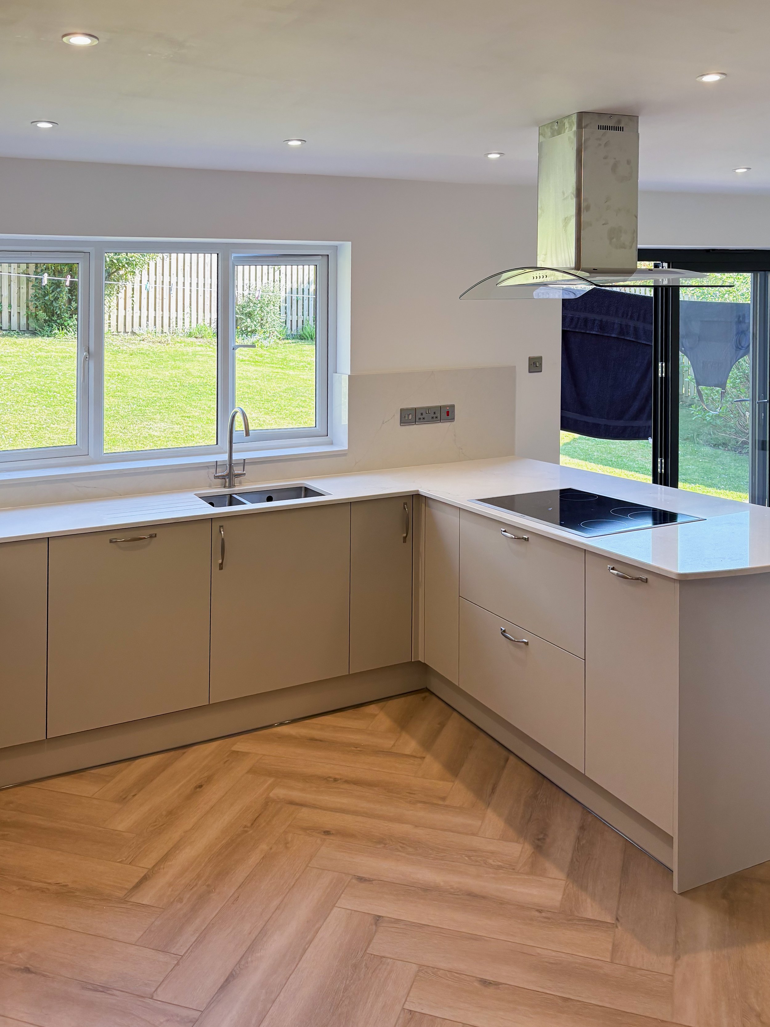 Modern kitchen with white countertops, beige cabinets, a window over the sink, and a stovetop with a stainless steel range hood, overlooking a grassy backyard.