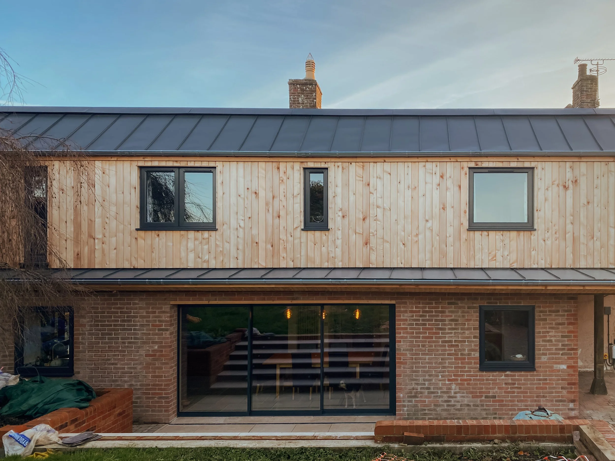 The rear view of a two-story house with brick ground floor and wooden upper floor, with large glass sliding doors and windows, a grey metal roof, and a chimney.