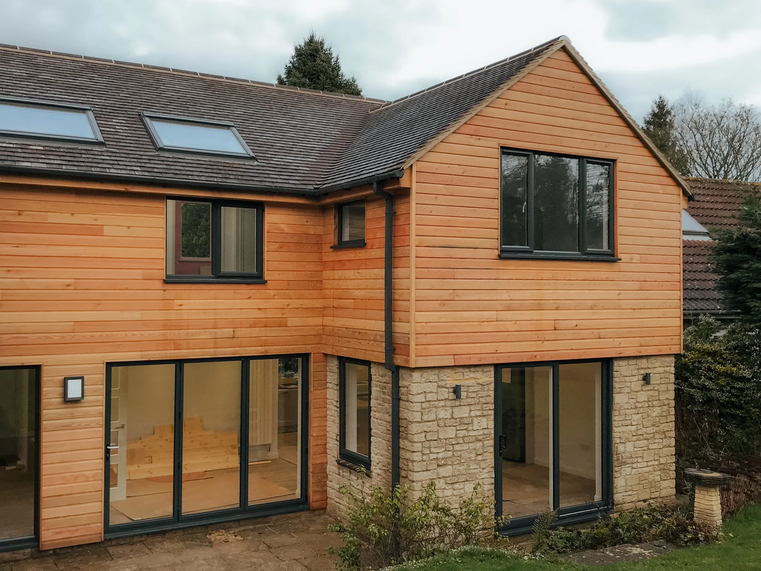 Modern two-story house with a combination of stone and wooden exterior, large glass sliding doors, and windows, with a sloped roof with skylights, surrounded by greenery.