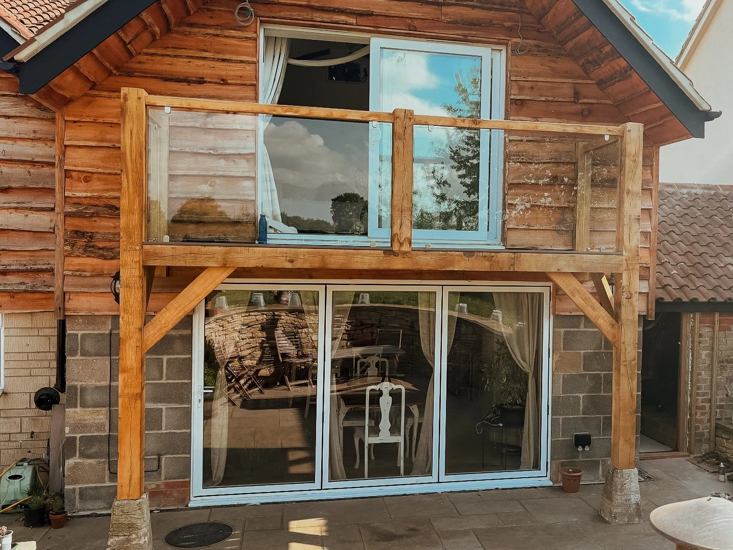 A two-story house with the upper level featuring a glass door and a small balcony with a glass railing. The house has a wooden exterior, large sliding glass doors on the ground level, and a tiled roof.