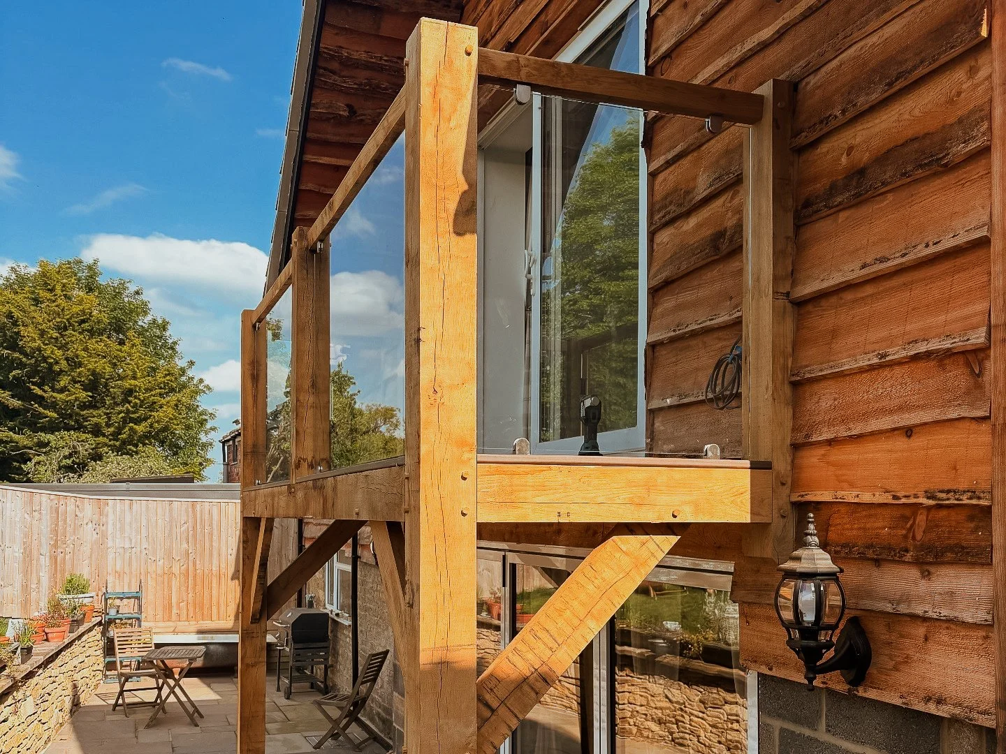 View of a wooden balcony under construction attached to a house with wooden siding, overlooking a backyard patio with chairs and a table, surrounded by a wooden fence, with a clear blue sky and trees in the background.