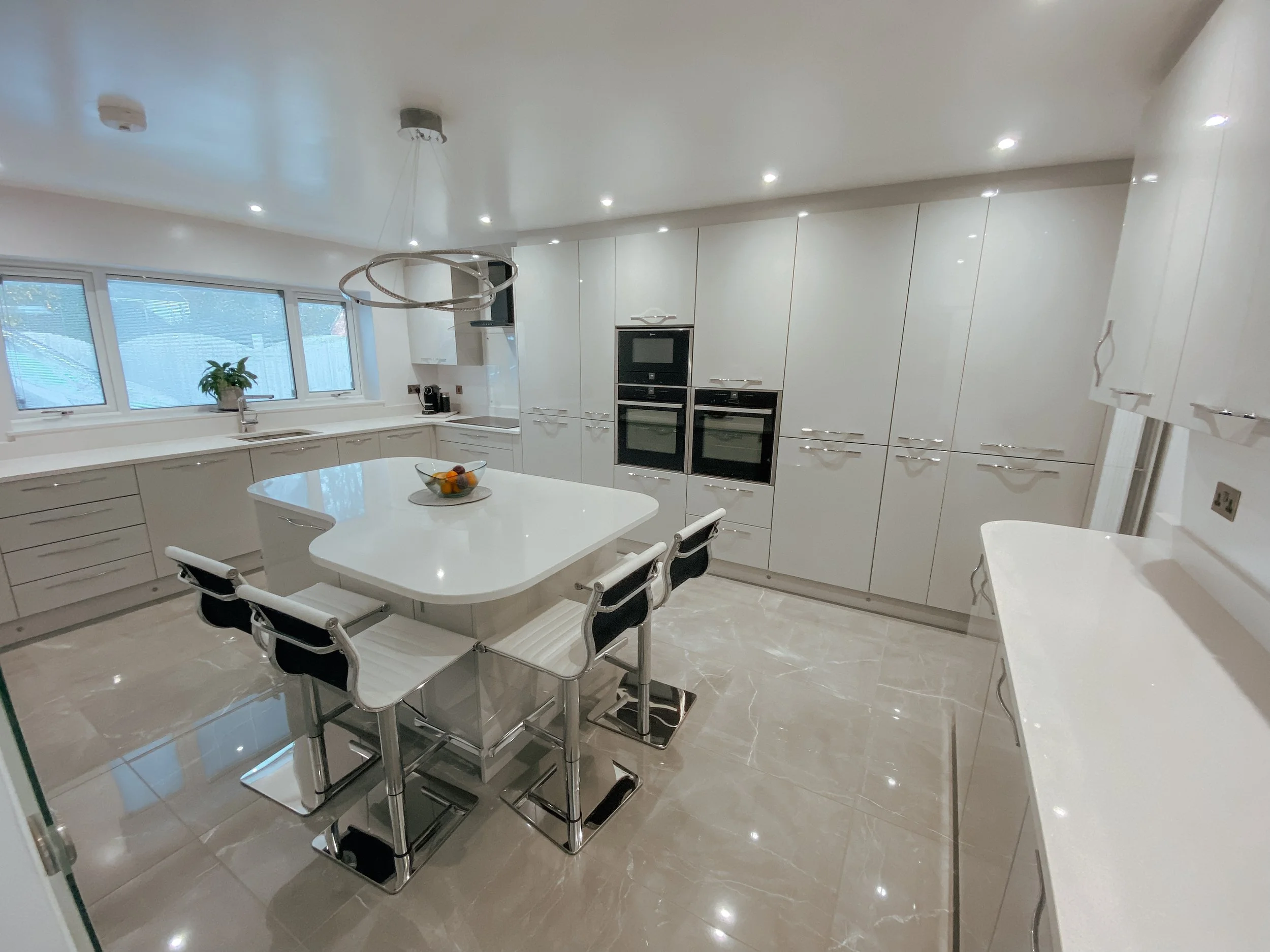 Modern white kitchen with built-in oven, microwave, and a central white dining table with black and white chairs. Contains a window with a plant, and a bowl of fruit on the table.