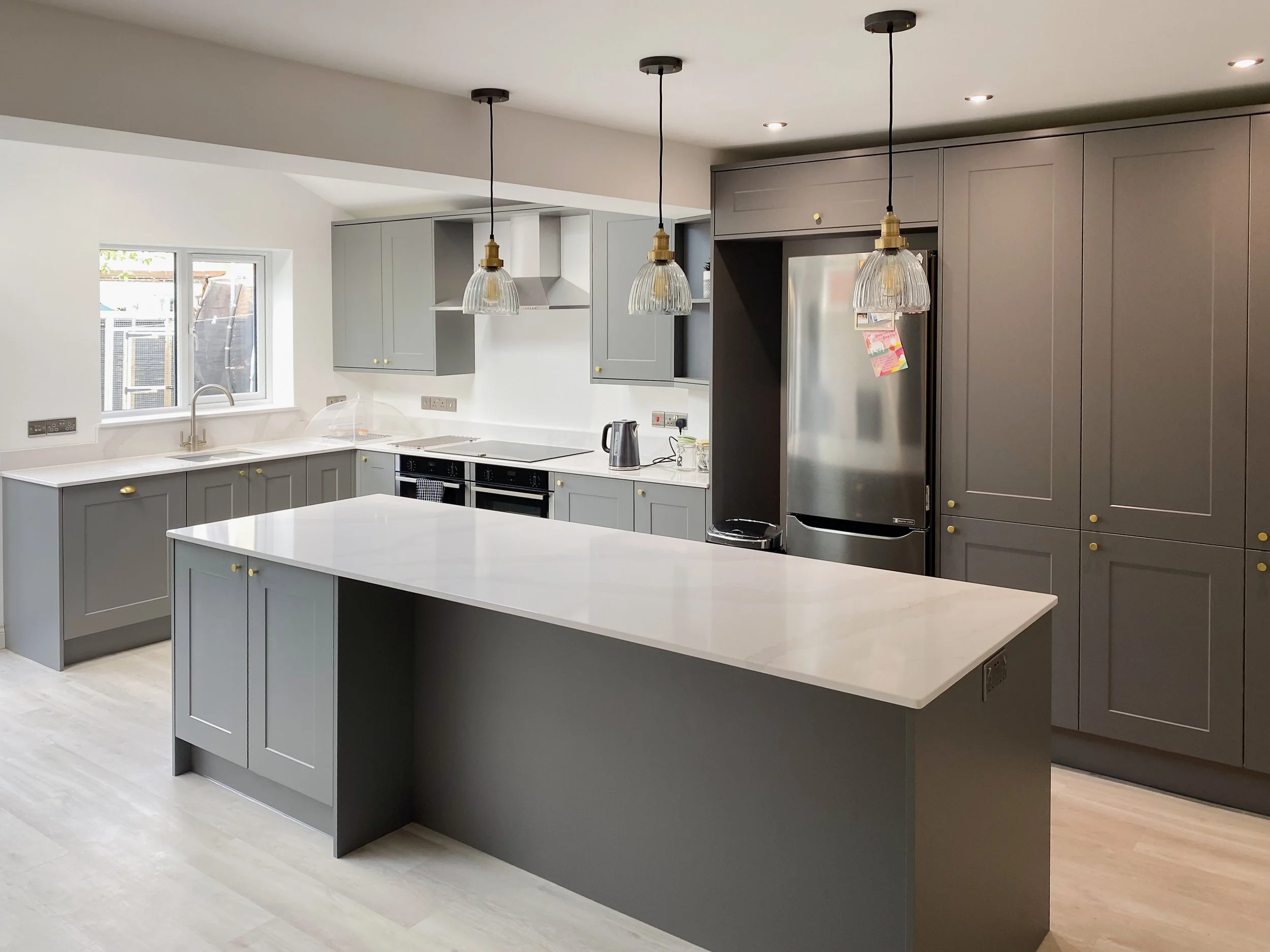 Modern kitchen with gray cabinets, white countertop island, stainless steel refrigerator, and pendant lights over the island.