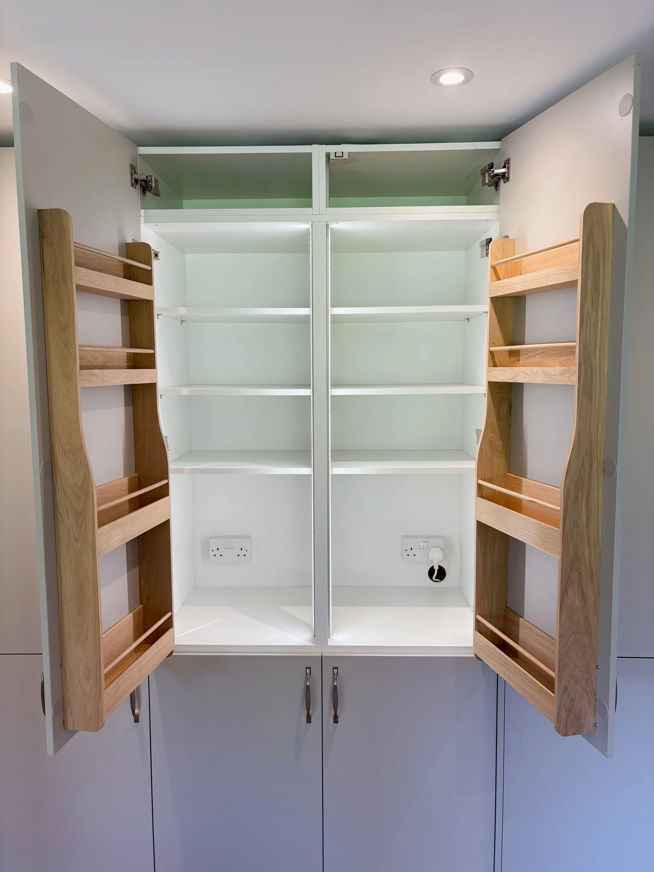 Empty white kitchen cabinet with open doors, featuring wooden spice rack organisers on the inside of each door and two electrical outlets inside the cabinet.