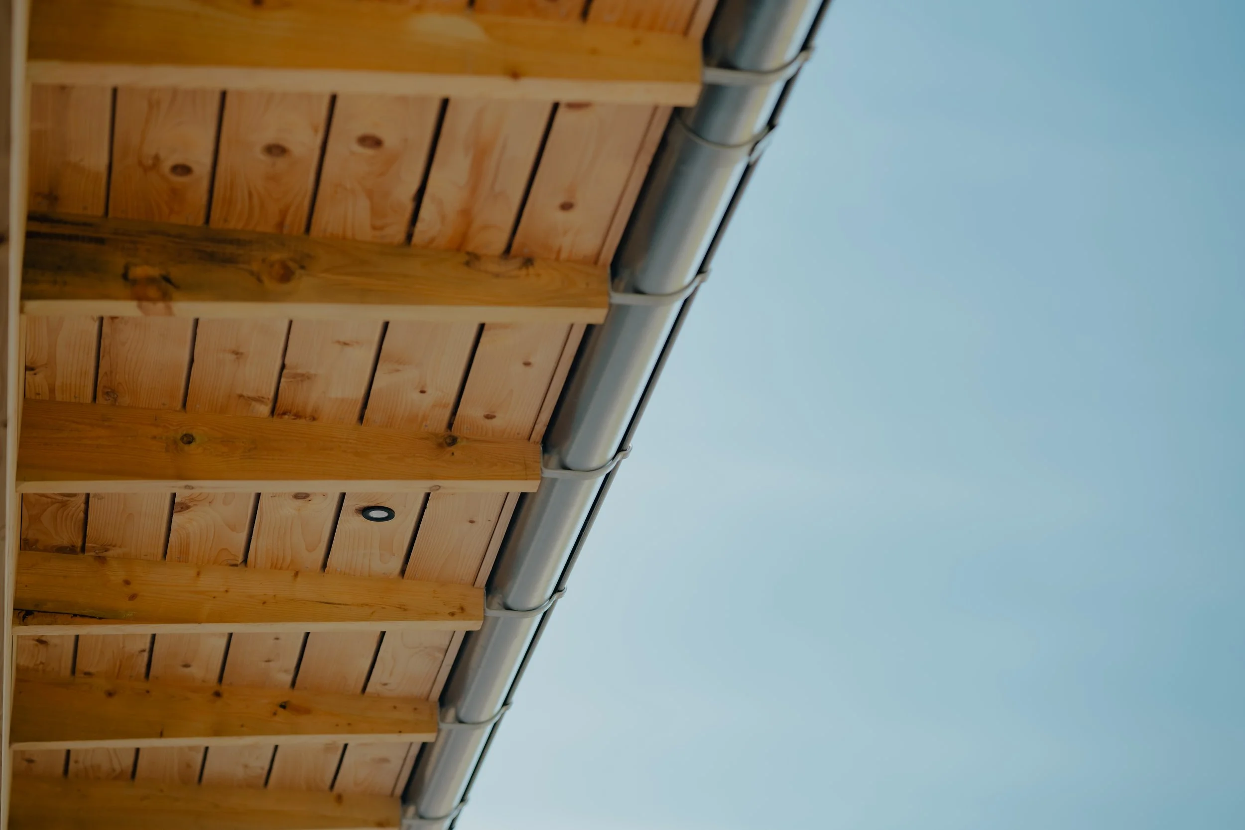 Close-up of a wooden roof structure under a clear blue sky, with a steel gutter along the edge.