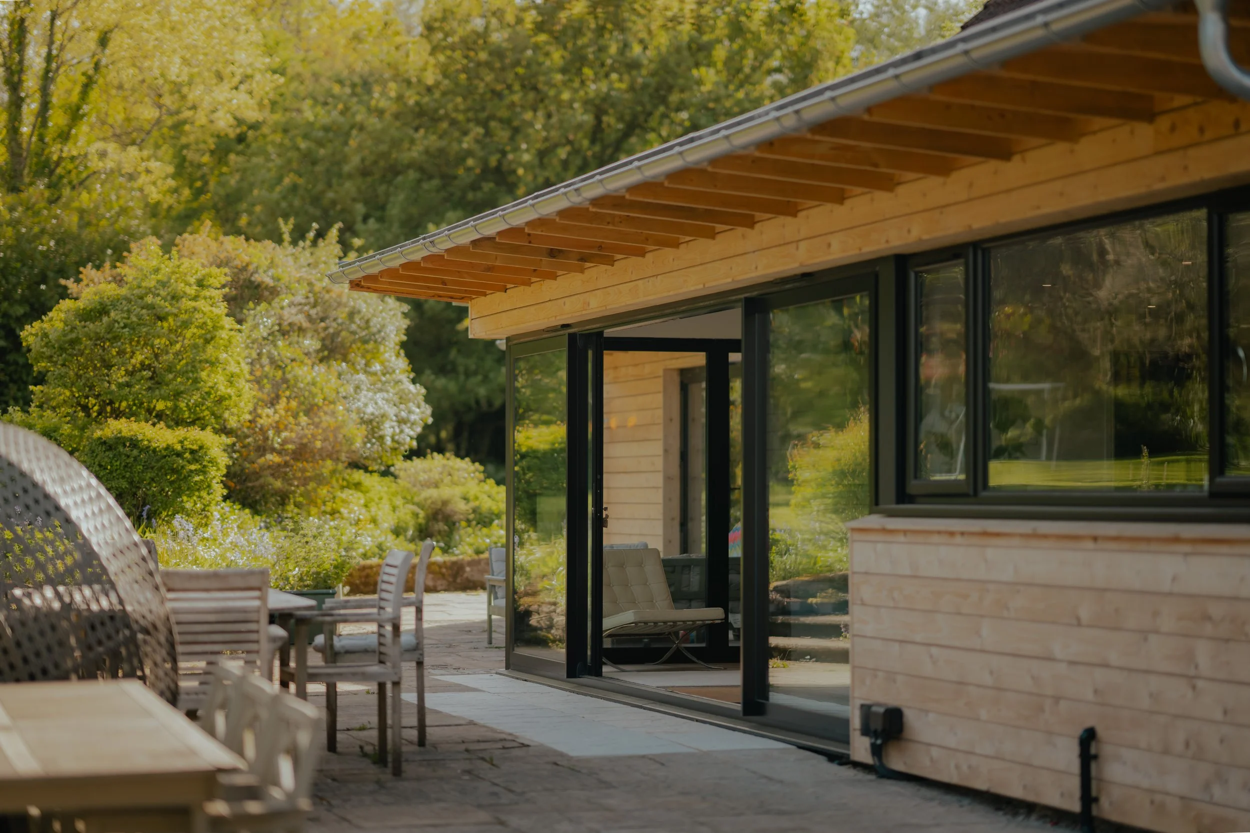Modern house with large glass sliding doors, wooden exterior, and patio furniture, surrounded by lush green trees in the background.