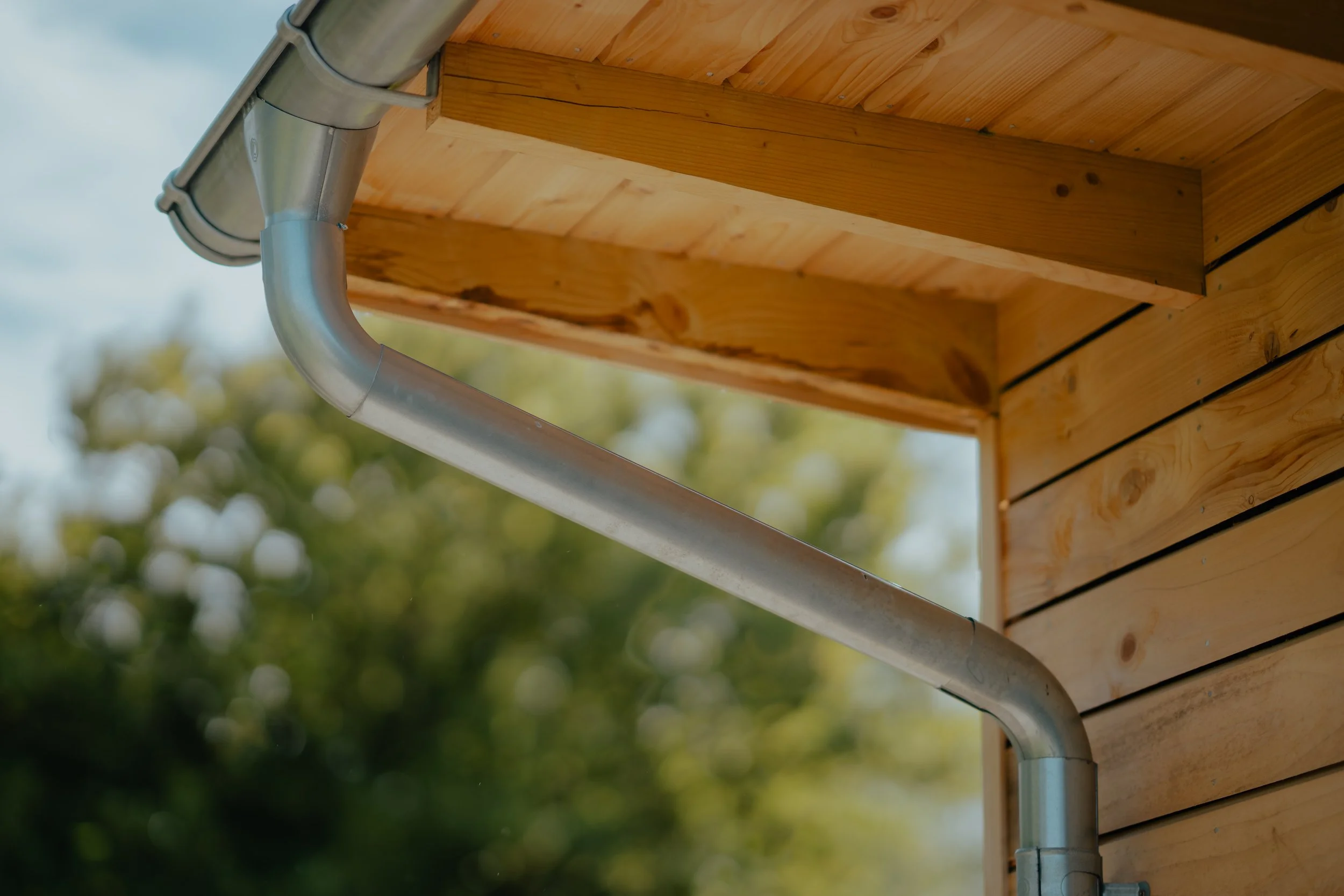 Close-up of a wooden roofing structure with a steel rain gutter attached, outdoors with blurred trees and sky in the background.