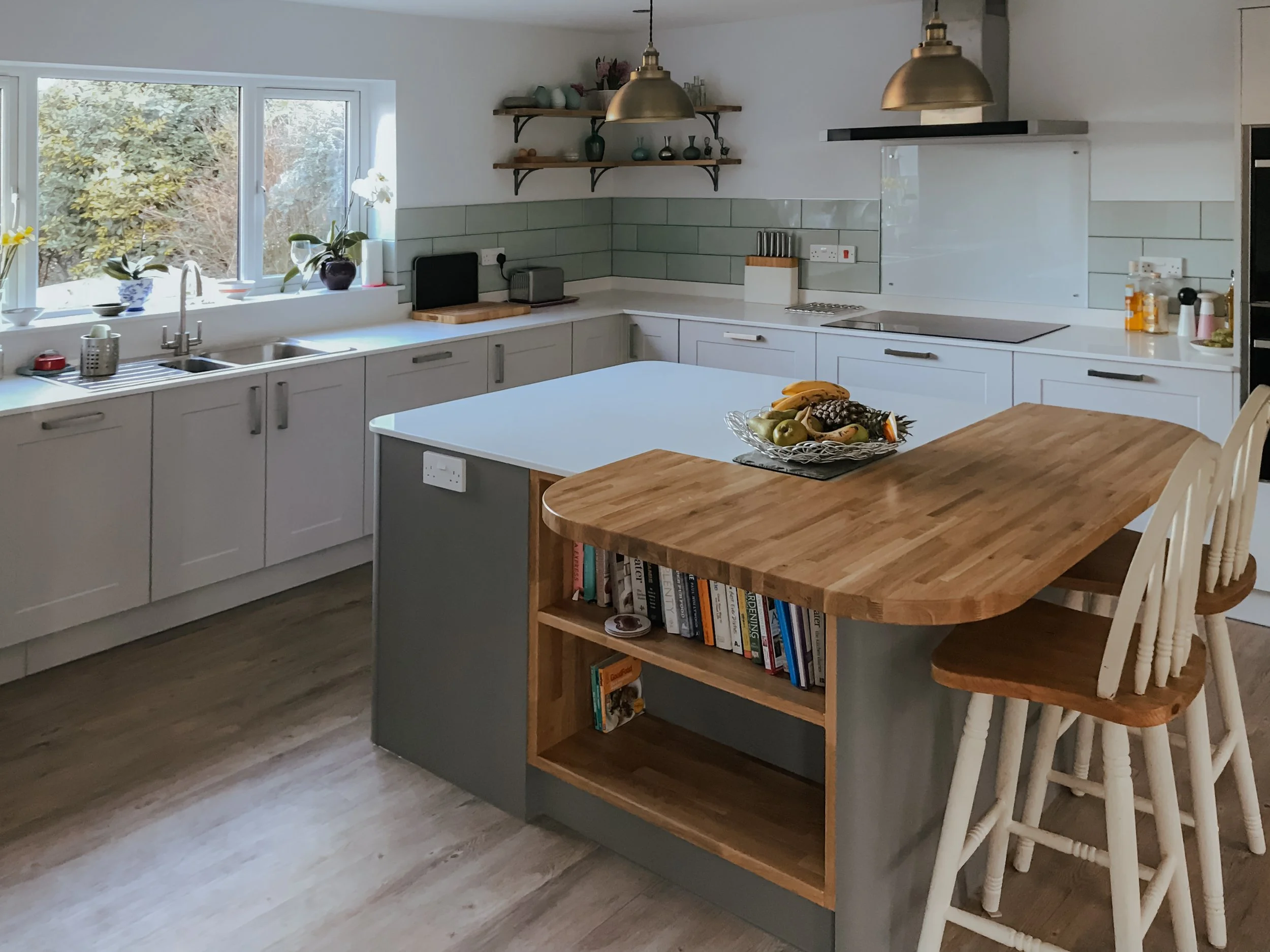 Bright, modern kitchen with white cabinets, a large window with plants, a wooden island with built-in bookshelf, and a fruit bowl on the island. There are barstools around the island and various appliances and decor on the counters.