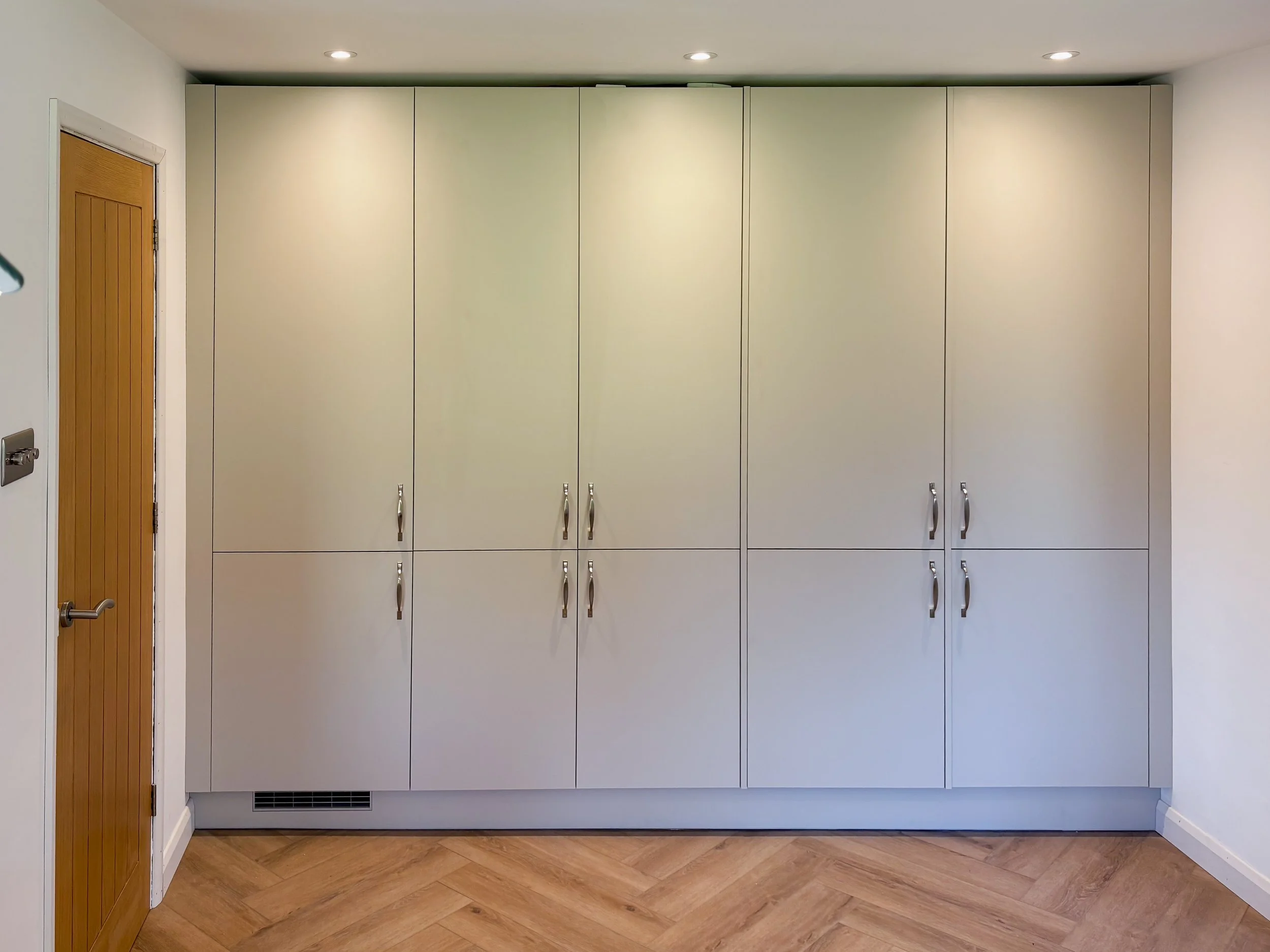 White storage cabinets with silver handles in a room, with a wooden door on the left and a hardwood floor.