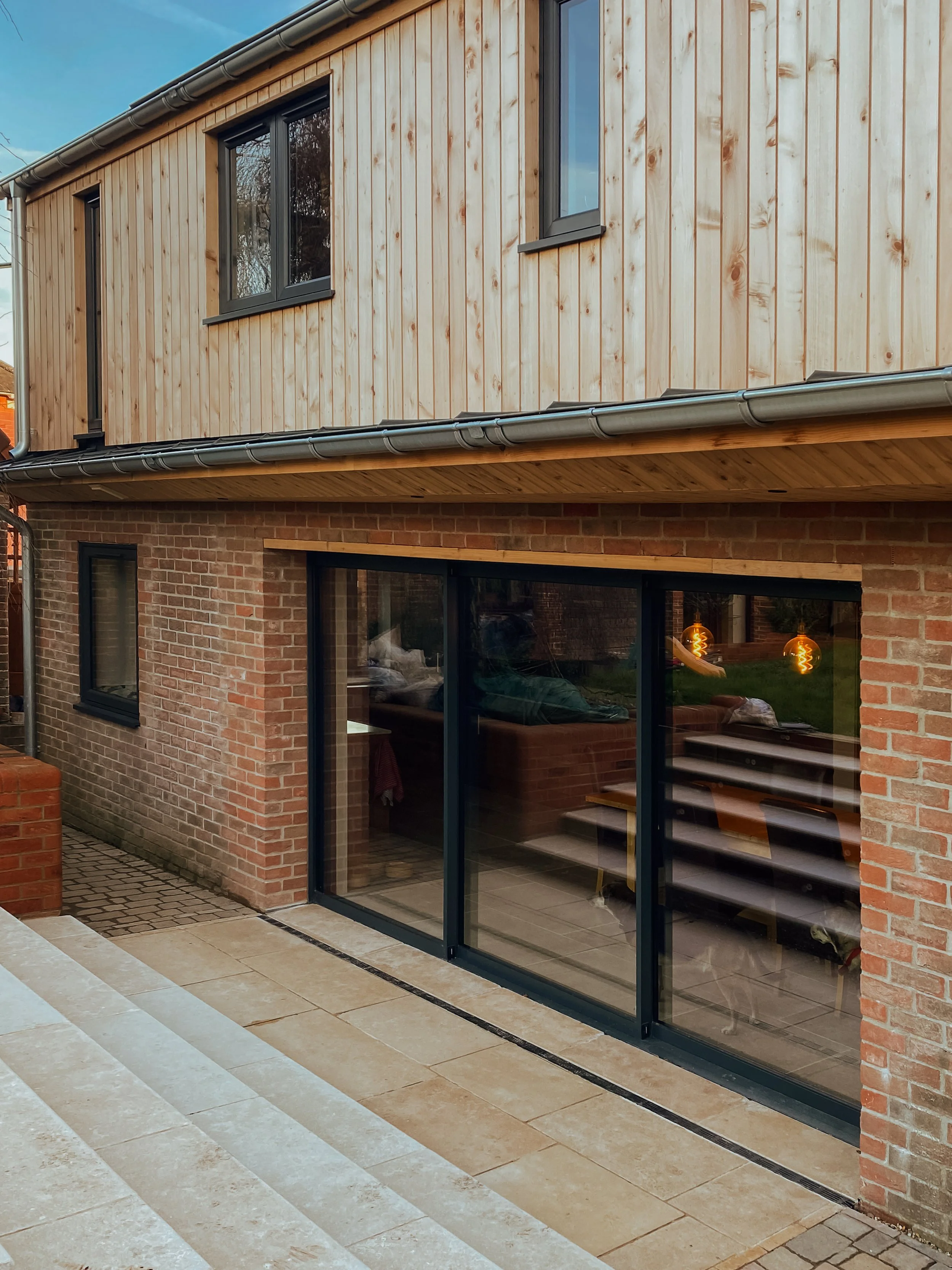 View of a two-story house with a brick ground level and wooden upper level, featuring large glass sliding doors and windows, with an outdoor paved area.