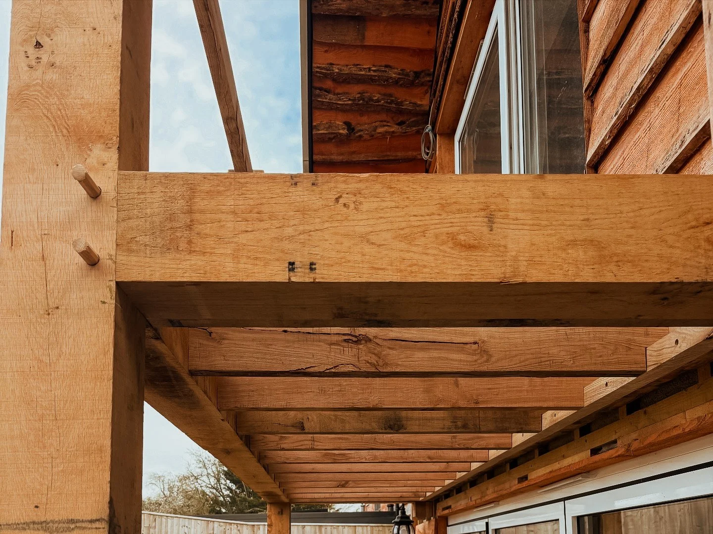 Construction of a wooden deck or balcony with exposed beams and support structure, overlooking a backyard with trees and a partly cloudy sky.