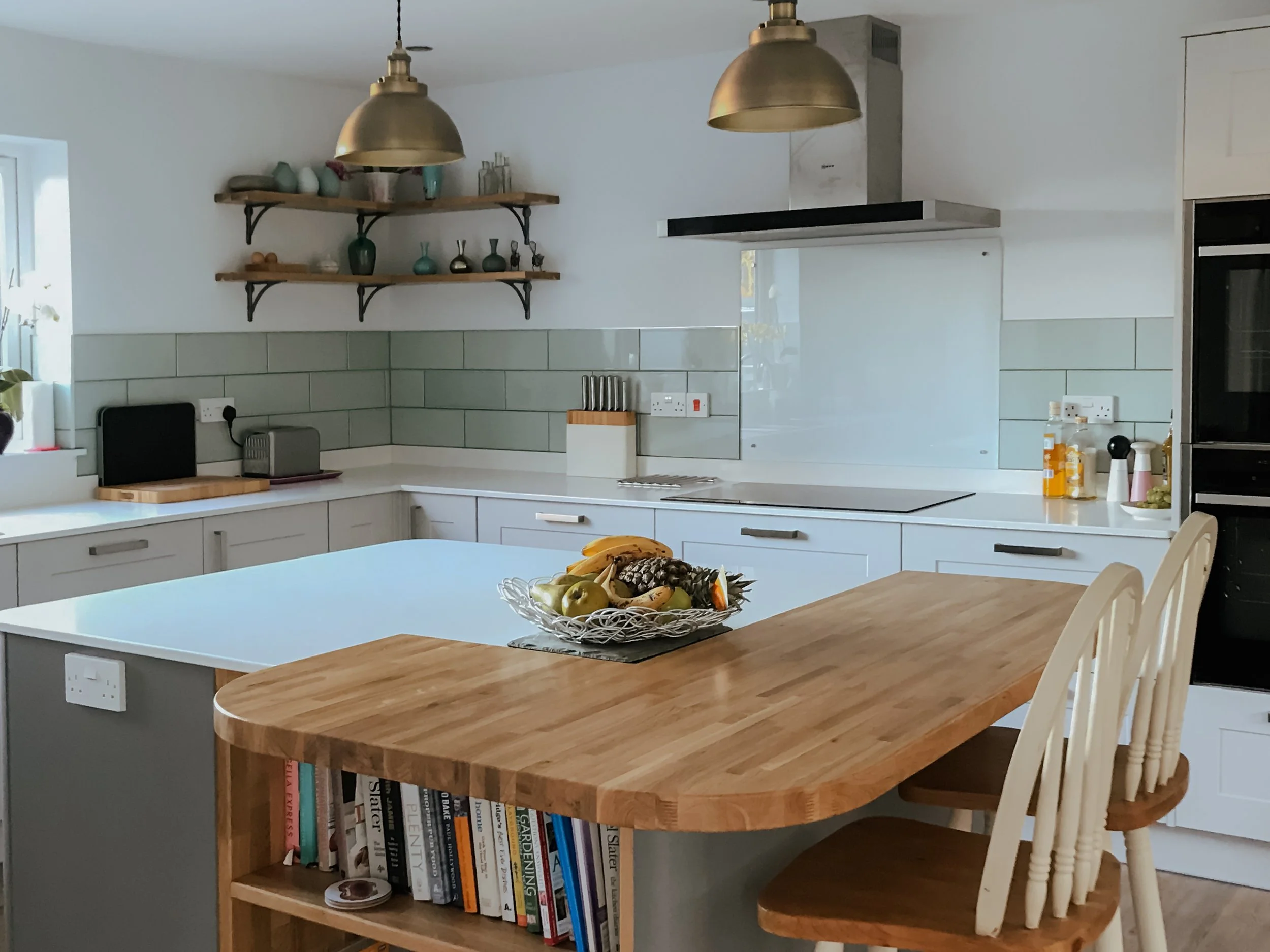 A modern kitchen with white cabinets, a white countertop, and sage green tiled backsplash. Open shelving with colorful vases and decorations. A fruit bowl with bananas, apples, and a pineapple on a wooden island. Hanging brass pendant lights, a built-in oven, and a window with a small plant. Books are stored on a shelf beneath the wooden countertop extending from the kitchen island.