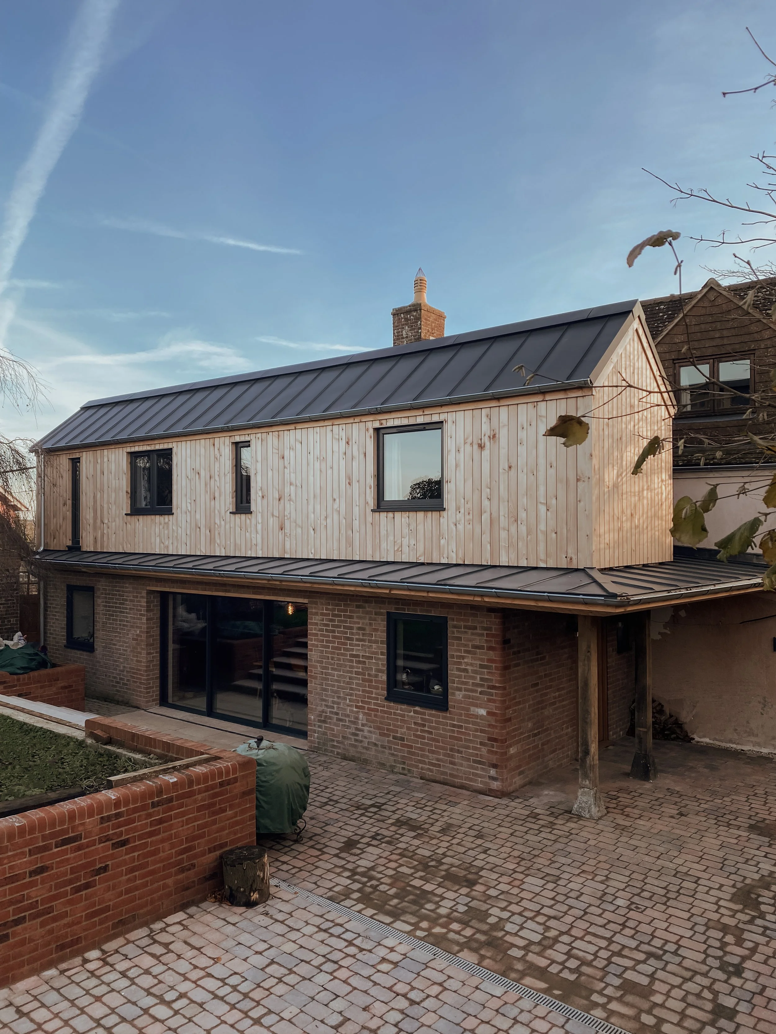 A modern two-story house with a brick lower level and a wooden upper level, large glass sliding doors, and multiple windows. The house features a metal roof and is situated in a yard with stone paving and brick walls.