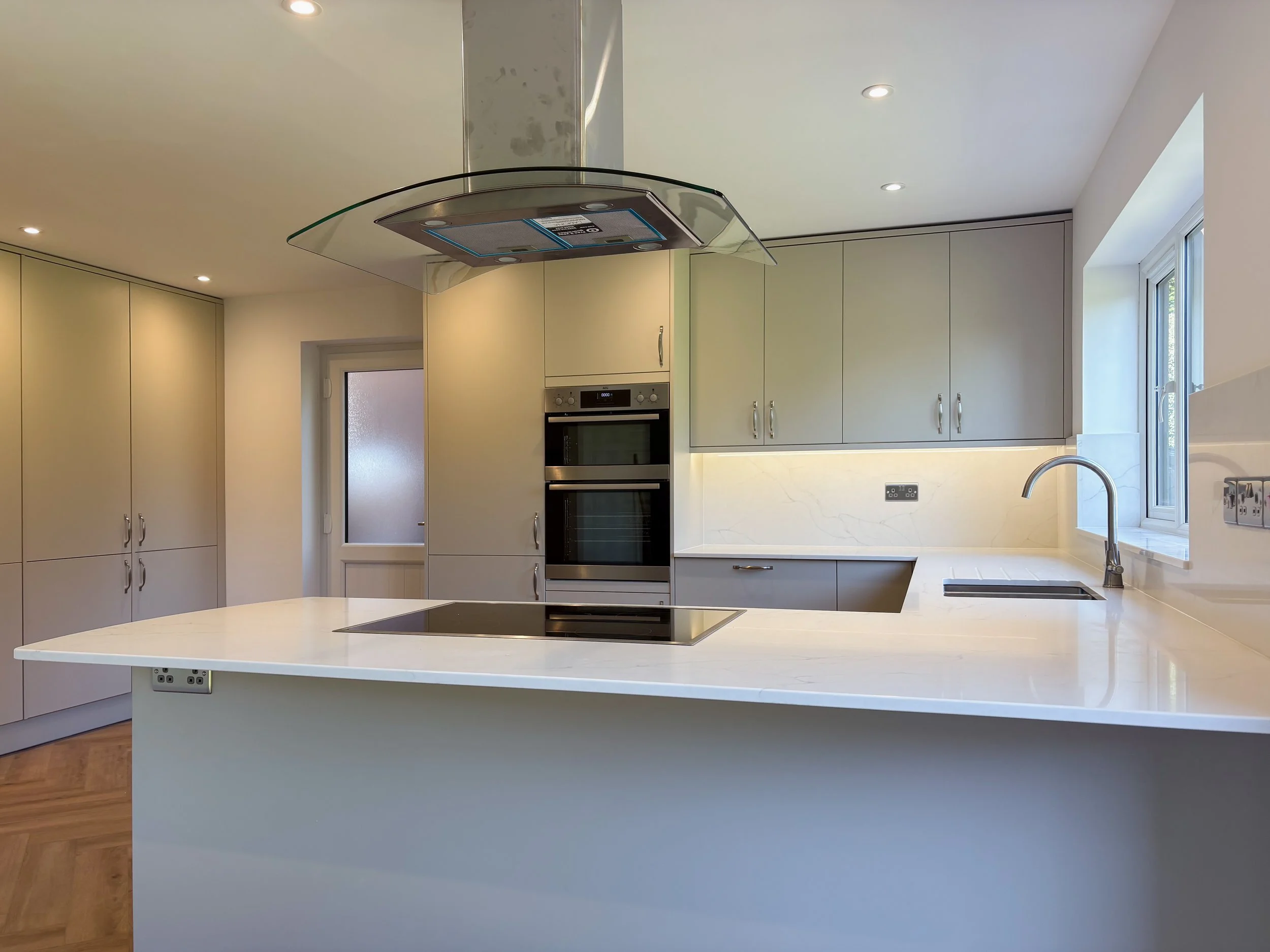 Modern kitchen with off-white cabinets, a white marble countertop island, a built-in oven, a black electric cooktop, a stainless steel range hood, a white marble backsplash, a window above the sink, and wood flooring.