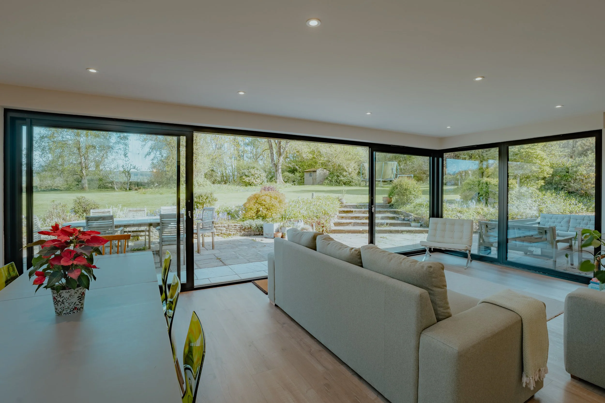 Living room with large glass sliding doors leading to a patio and backyard with outdoor furniture, trees, and a shed, furnished with a white sofa, glass-top dining table with poinsettia plant, and other seating.