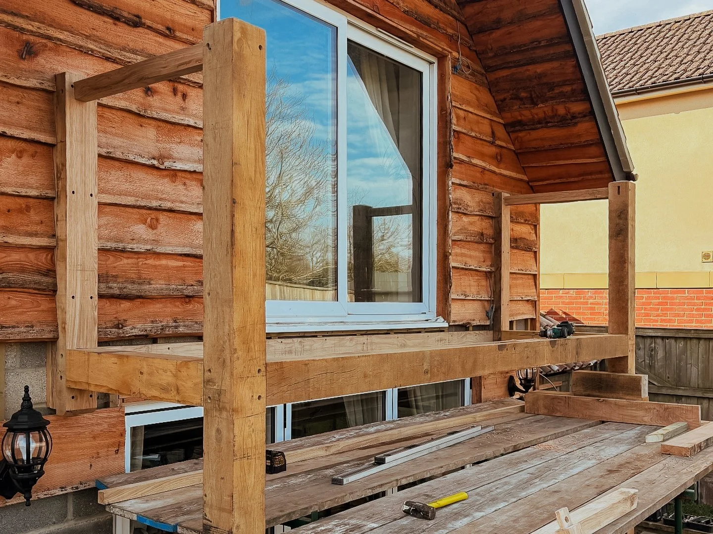 Wooden deck railing under construction outside a house with wooden siding and large window.