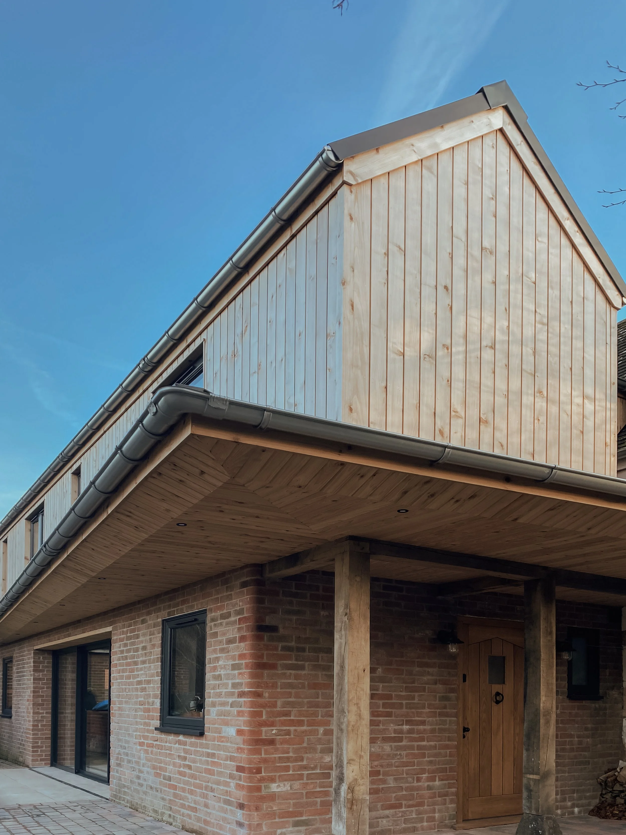 A two-story house with a brick lower level and wooden upper level, featuring a sloped roof, black-framed windows, and a wooden front door under a covered porch. The sky is blue.