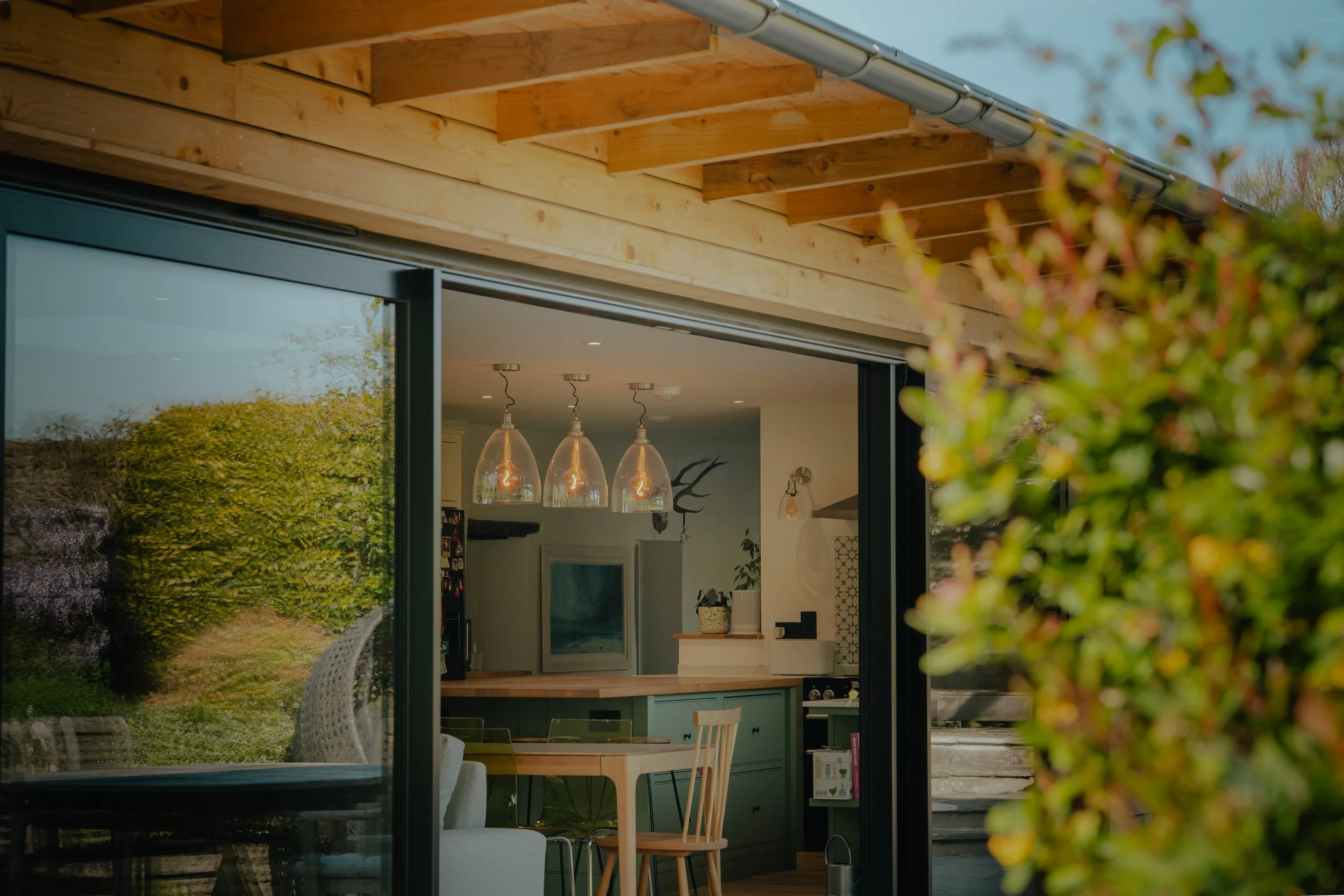 View of a house interior through a large open sliding glass door, showing a kitchen with hanging pendant lights, a dining table with chairs, and indoor plants, with a garden and trees outside.