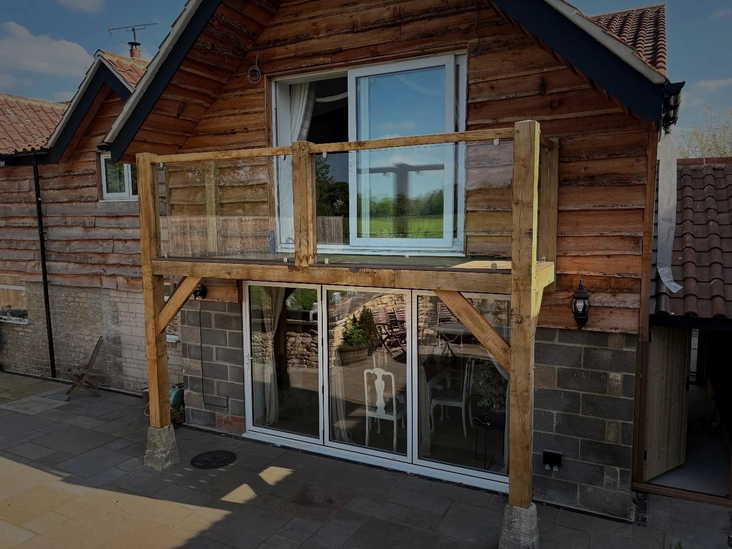 A two-story house with a wooden exterior, a glass sliding door on the ground floor, and a window on the upper floor. There is wooden balcony with a railing above the ground level, supported by wooden beams, with a sliding glass door leading to the balcony.