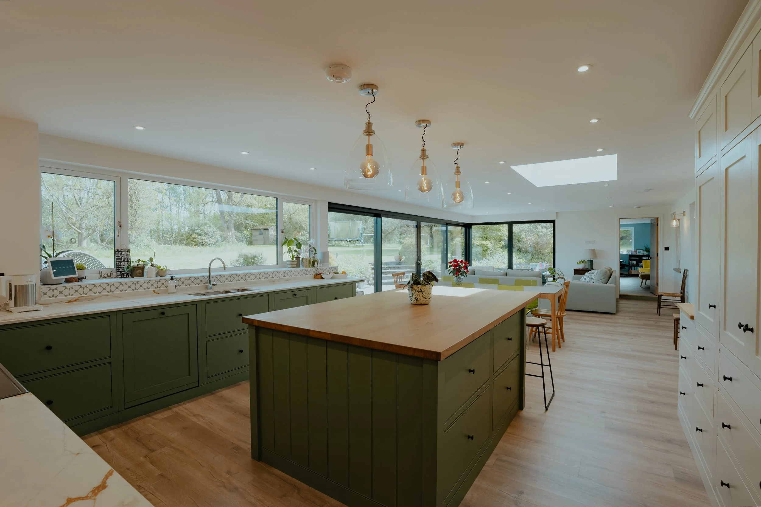 Open-concept kitchen and living room with large windows, a wooden island with green cabinetry, pendant lights, and modern decor.