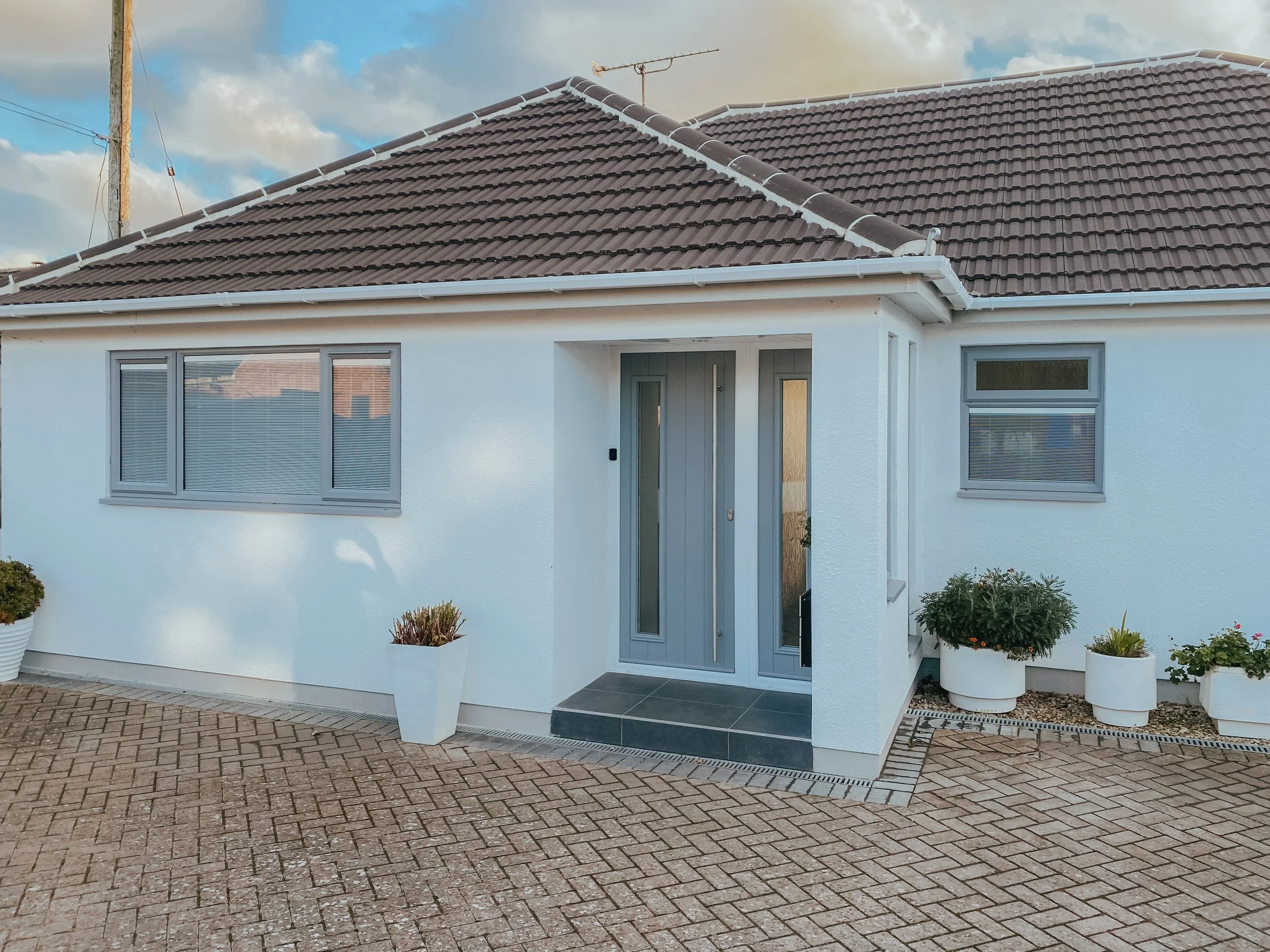 Front view of a white house with a brown tiled roof, gray window frames, and a gray front door. There are potted plants on either side of the entrance and a paved driveway in front.