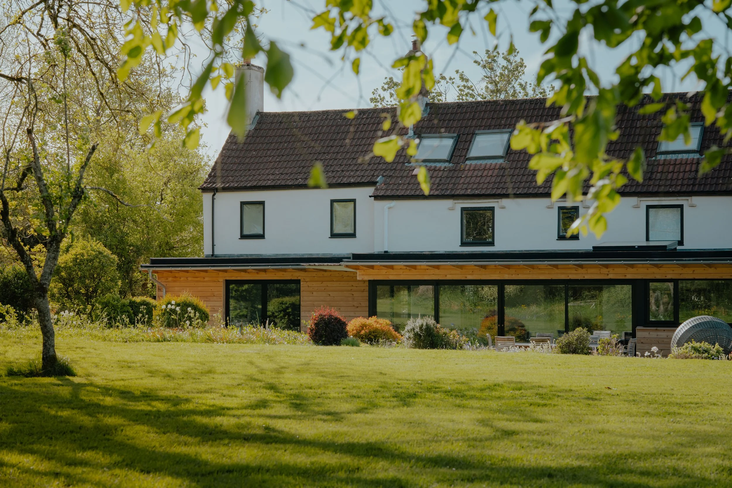 A modern house with white walls, a dark tiled roof, and large windows, surrounded by a lush green garden with trees and flowering shrubs, under a clear blue sky.