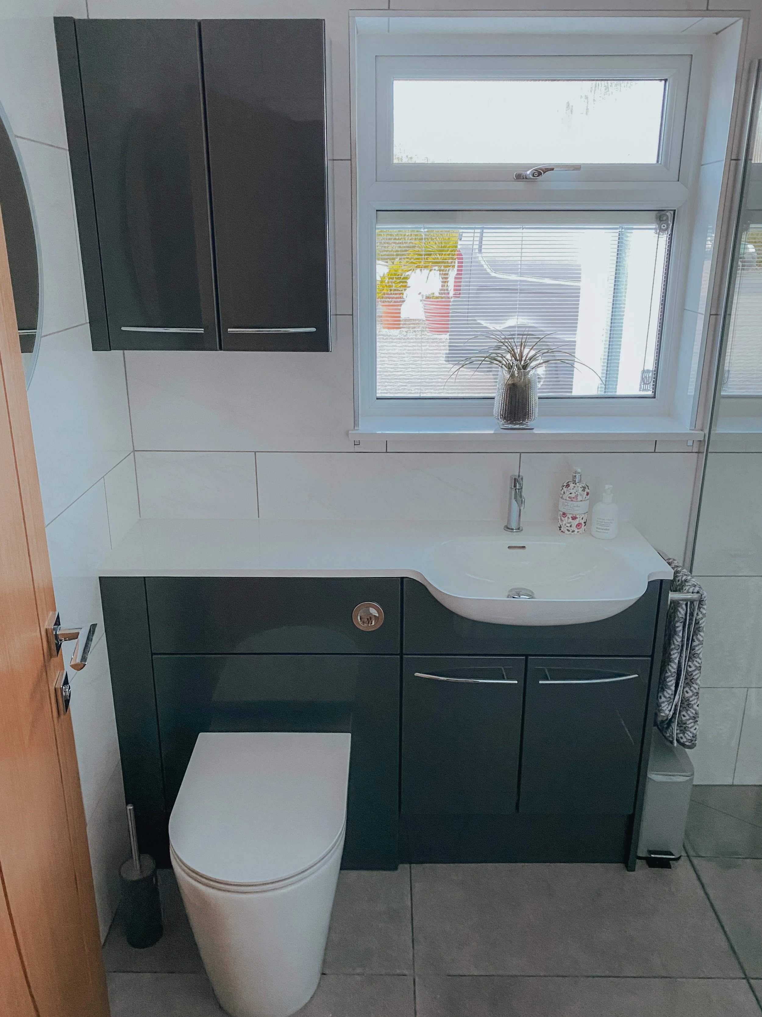 A bathroom with white tiled walls, a window with a potted plant on the windowsill, dark green cabinets below and above the sink, a white toilet to the left of the sink, and a small trash bin to the right of the toilet.