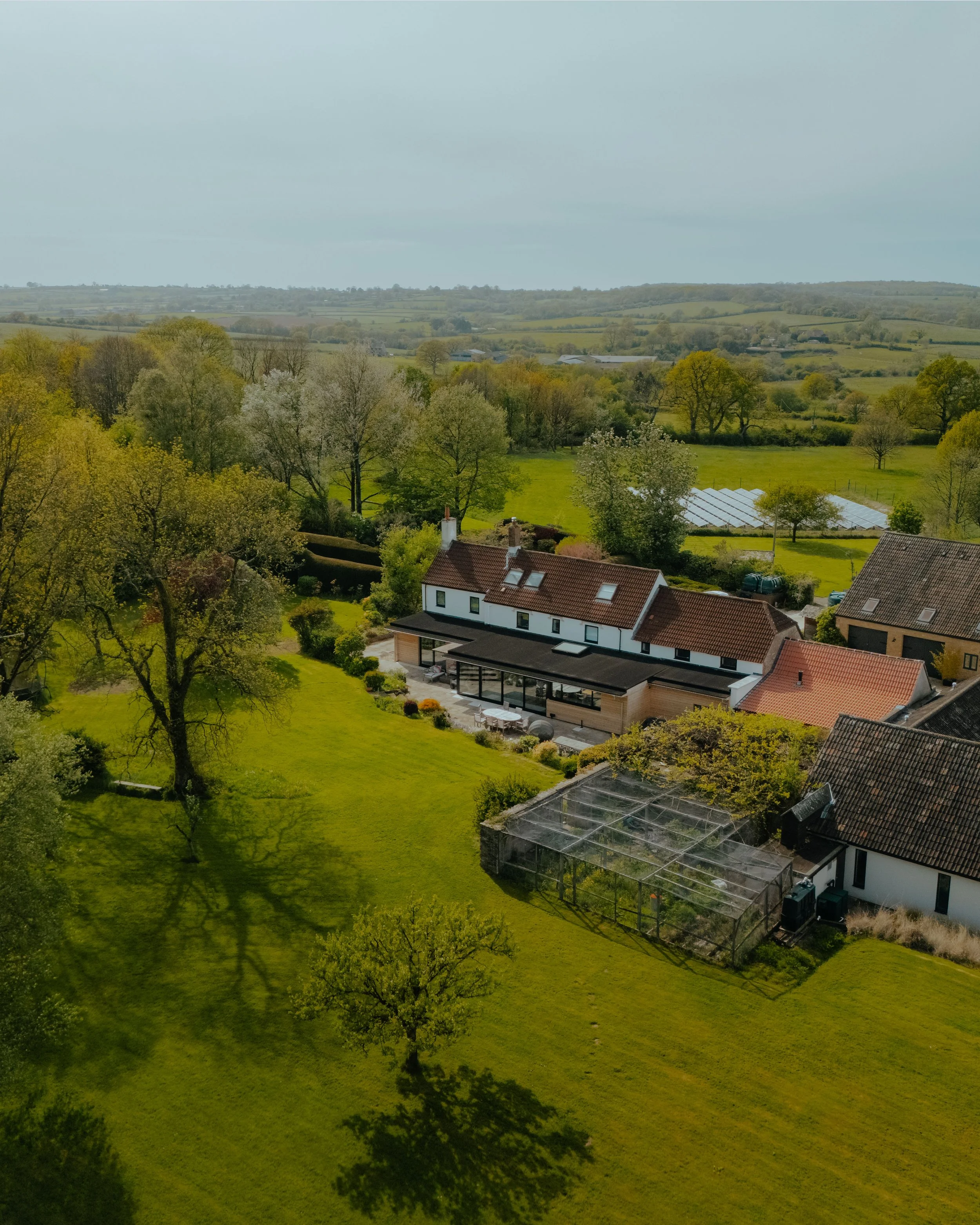 A peaceful rural landscape with a mix of deciduous trees, green fields, and the contemporary home extension and renovation, including a house with a patio and a greenhouse, under a partly cloudy sky.