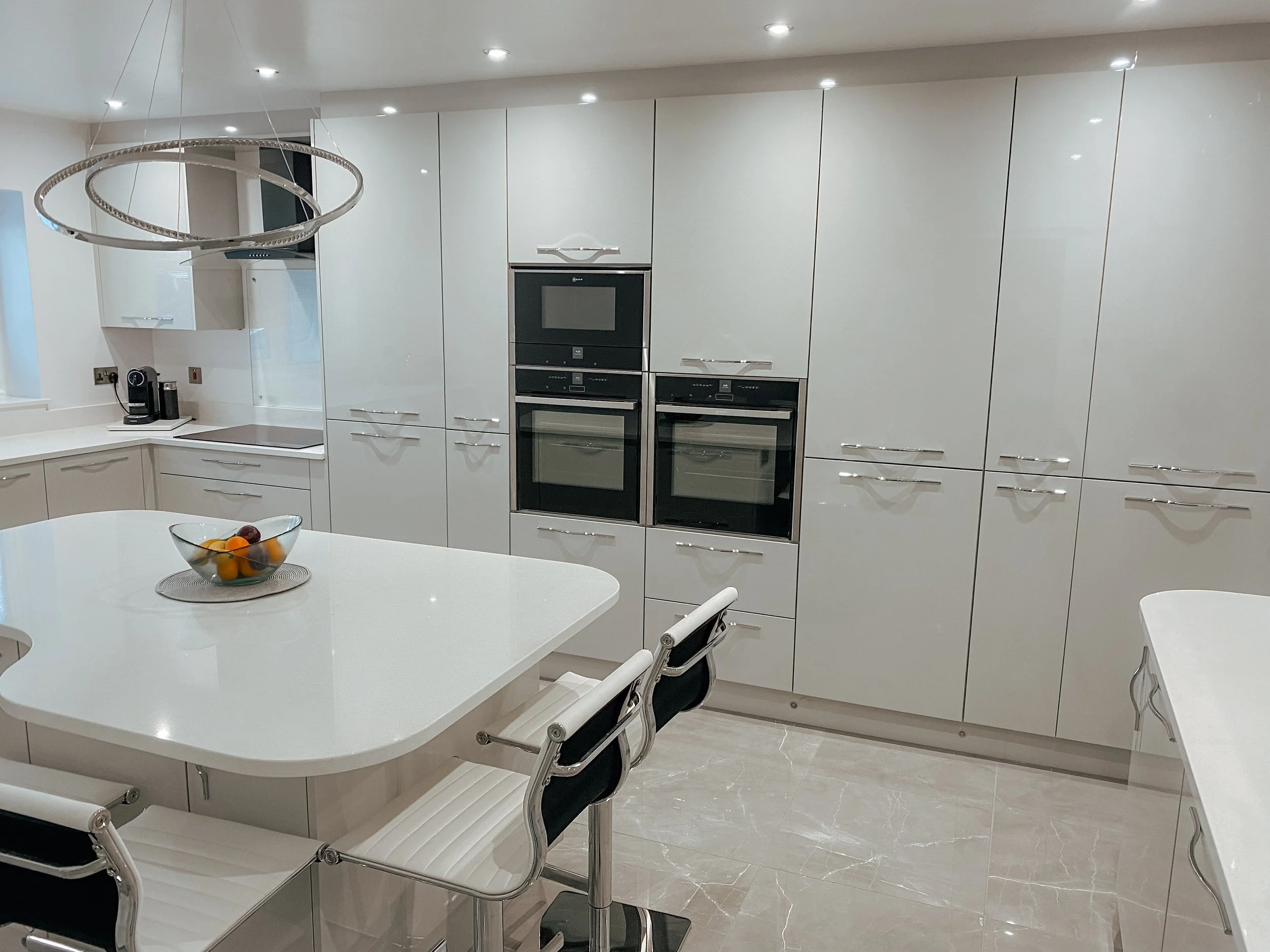 Modern white kitchen with built-in double oven, small table with chairs, and a fruit bowl.