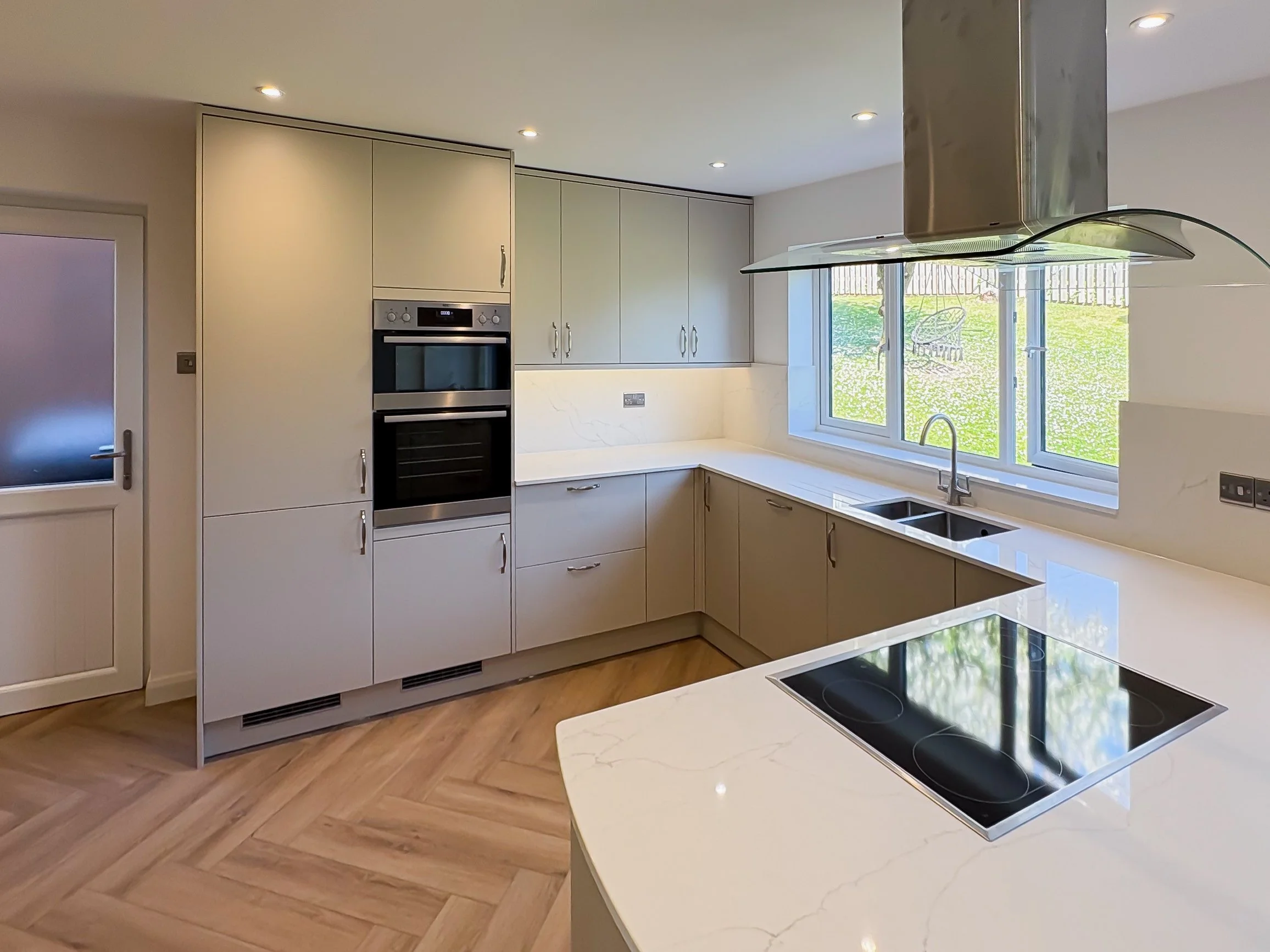 Modern kitchen with beige and white cabinetry, marble countertops, a built-in oven, a stovetop on the island, a window overlooking a grassy yard, and a stainless steel range hood.
