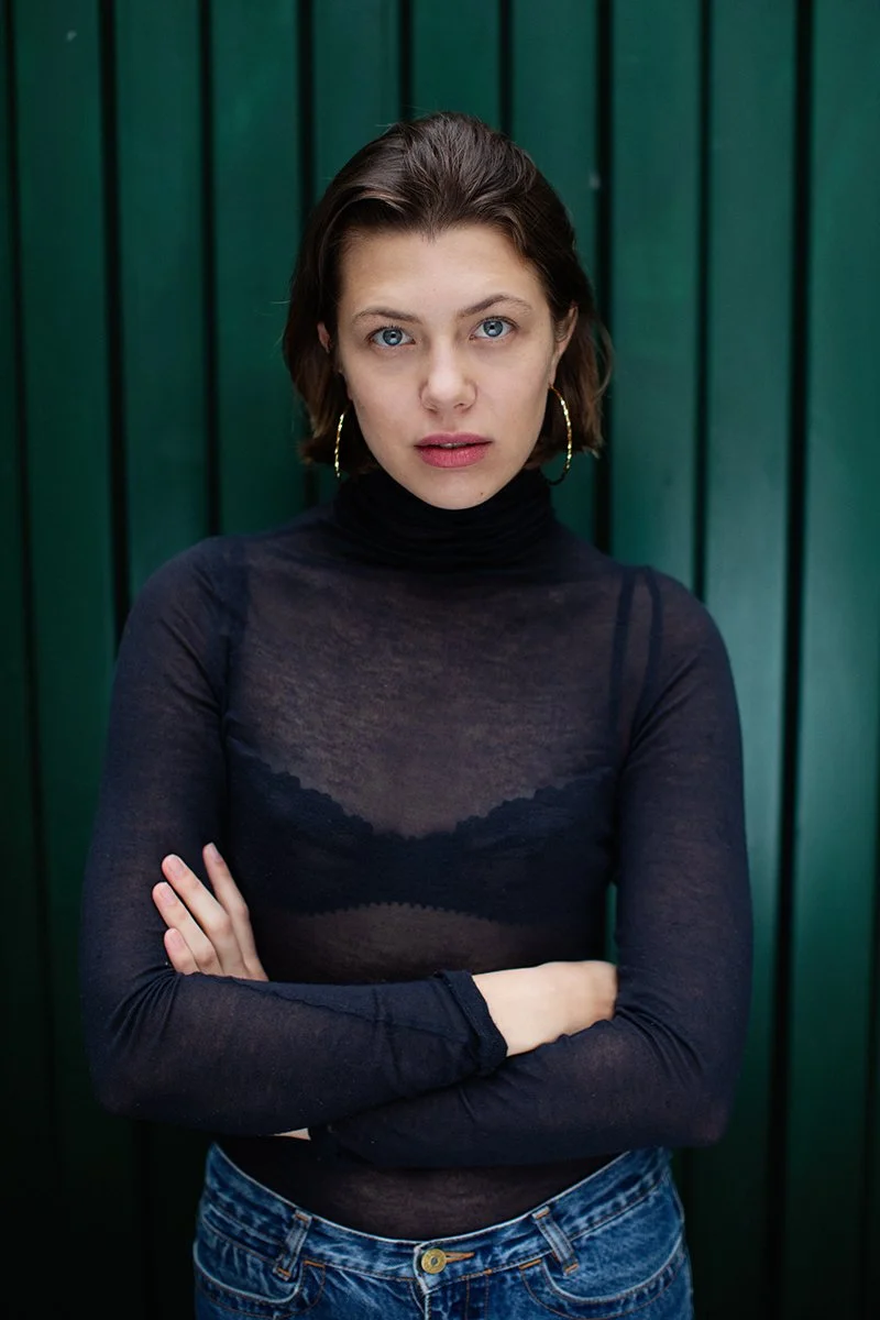 Eine junge Frau mit kurzen braunen Haaren, blauen Augen, schwarzen langen Arm- und Schulterfreiem Oberteil und Jeans steht vor einer grünen Holzwand, die Arme verschränkt.