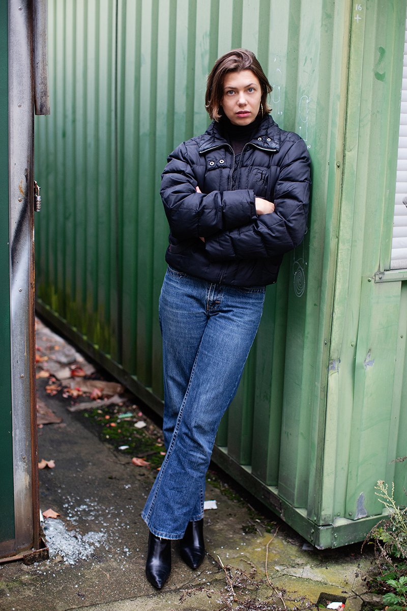 Eine junge Frau mit kurzen braunen Haaren, dunklem Pullover und schwarzer Daunenjacke, steht mit verschränkten Armen vor einer grünen Metallwand im Freien.