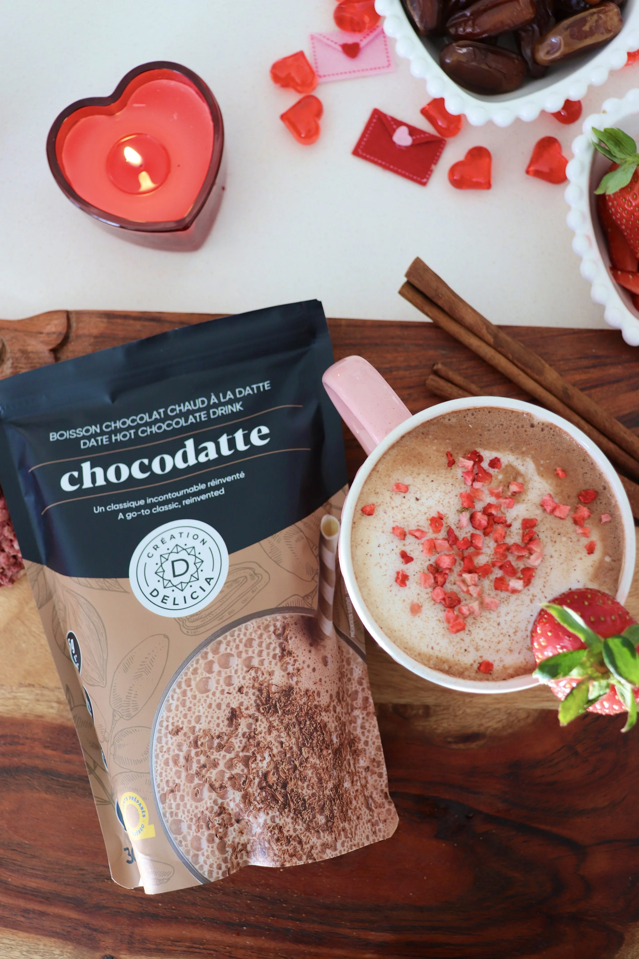 Une tasse de chocolat chaud à la vanille avec des morceaux de fraise et des morceaux de sucre rose, une poudre de cacao dans un paquet, et une petite bougie en forme de cœur sur une table en bois, entourée de bonbons, de fraises et d'autres éléments décoratifs.