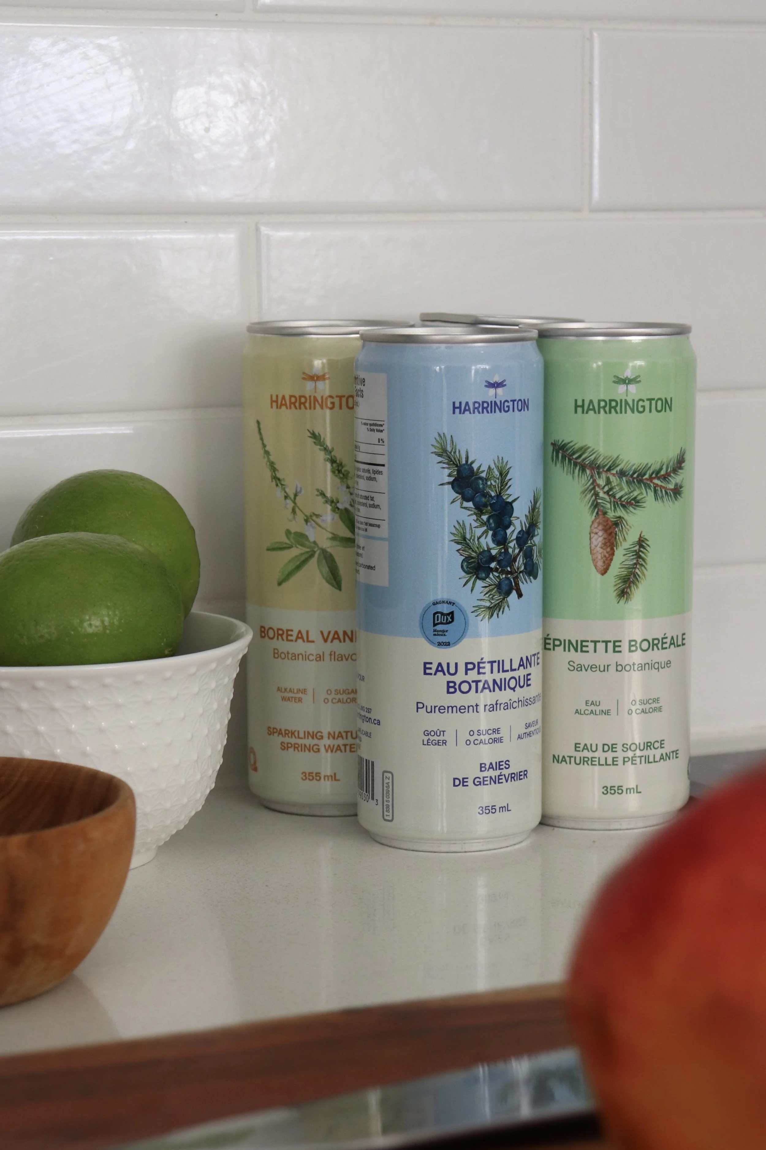 Trois canettes d'eau gazeuse botanique de Harrington, un bol blanc avec deux citrons verts, et une partie d'une pomme rouge en premier plan sur une étagère blanche en arrière-plan.