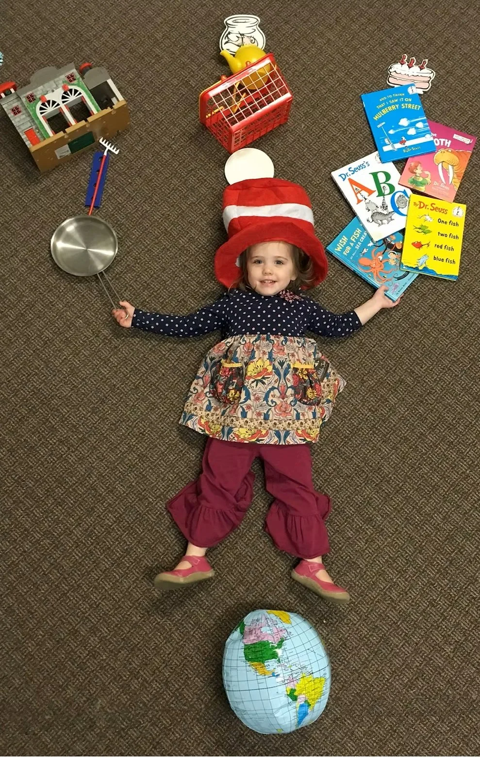 A young girl lying on a brown carpet, wearing a red and white hat that resembles the Dr. Seuss character, The Cat in the Hat. She is surrounded by children's books, a toy shopping cart, a toy house, a metal pan, and a plush globe.