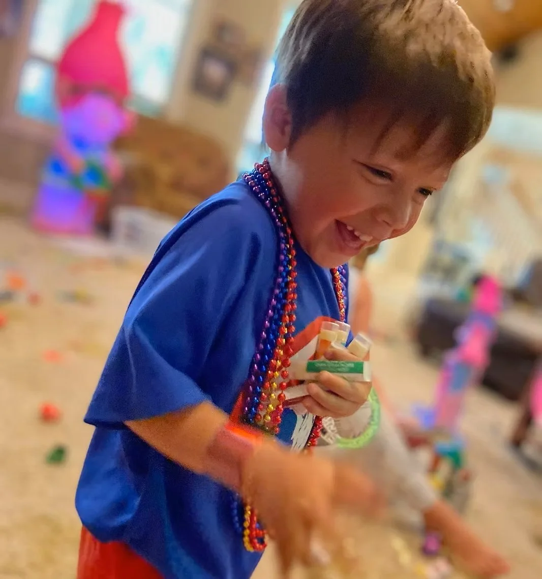 A joyful young boy wearing multicolored bead necklaces and a blue shirt, holding small sandwiches, amidst a lively indoor celebration with scattered confetti.