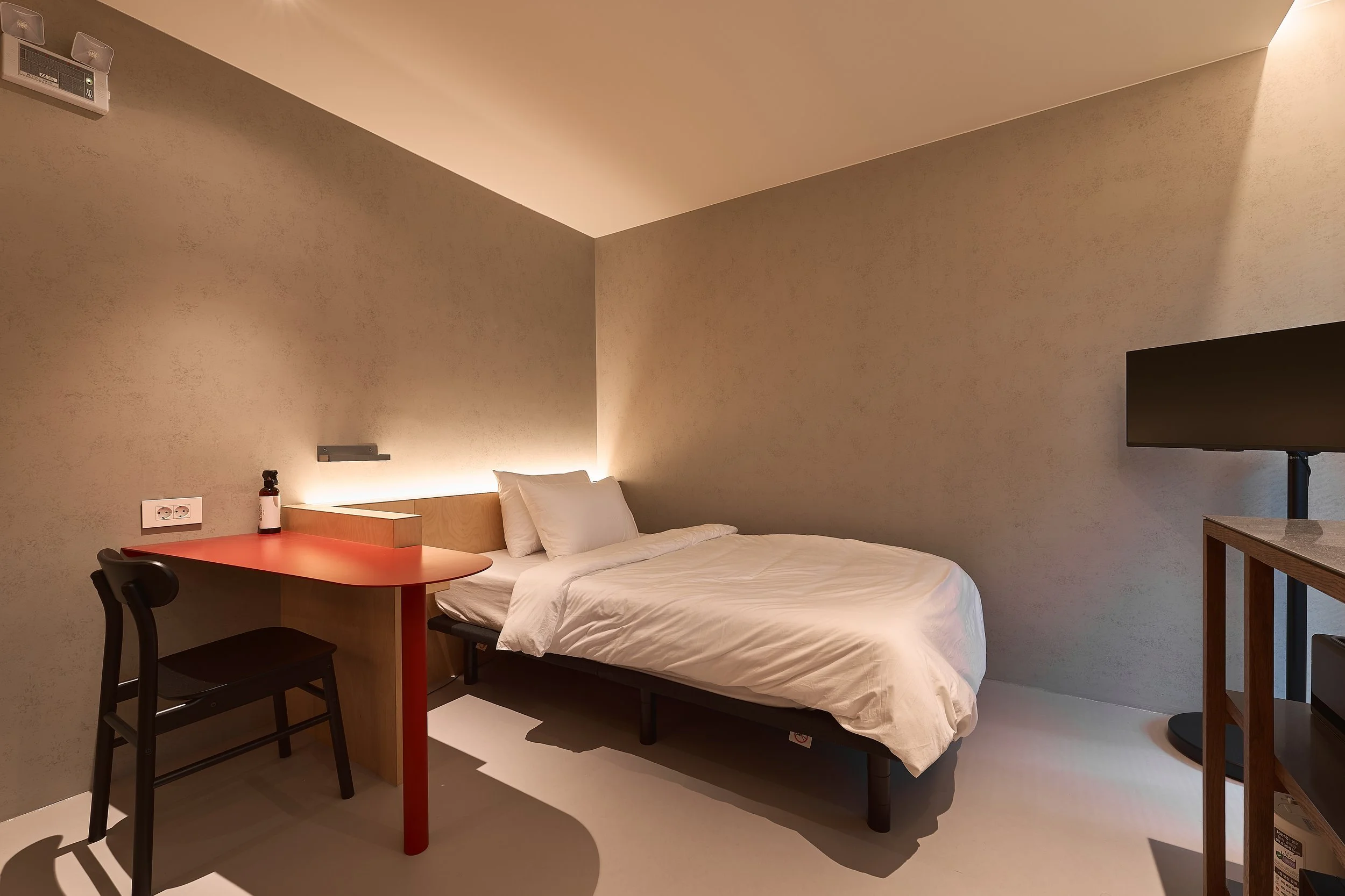 willow leaf Gangnam Room 201 featuring a minimalist bedroom with a cozy bed, warm indirect lighting, red work desk, black chair, and wall-mounted TV.