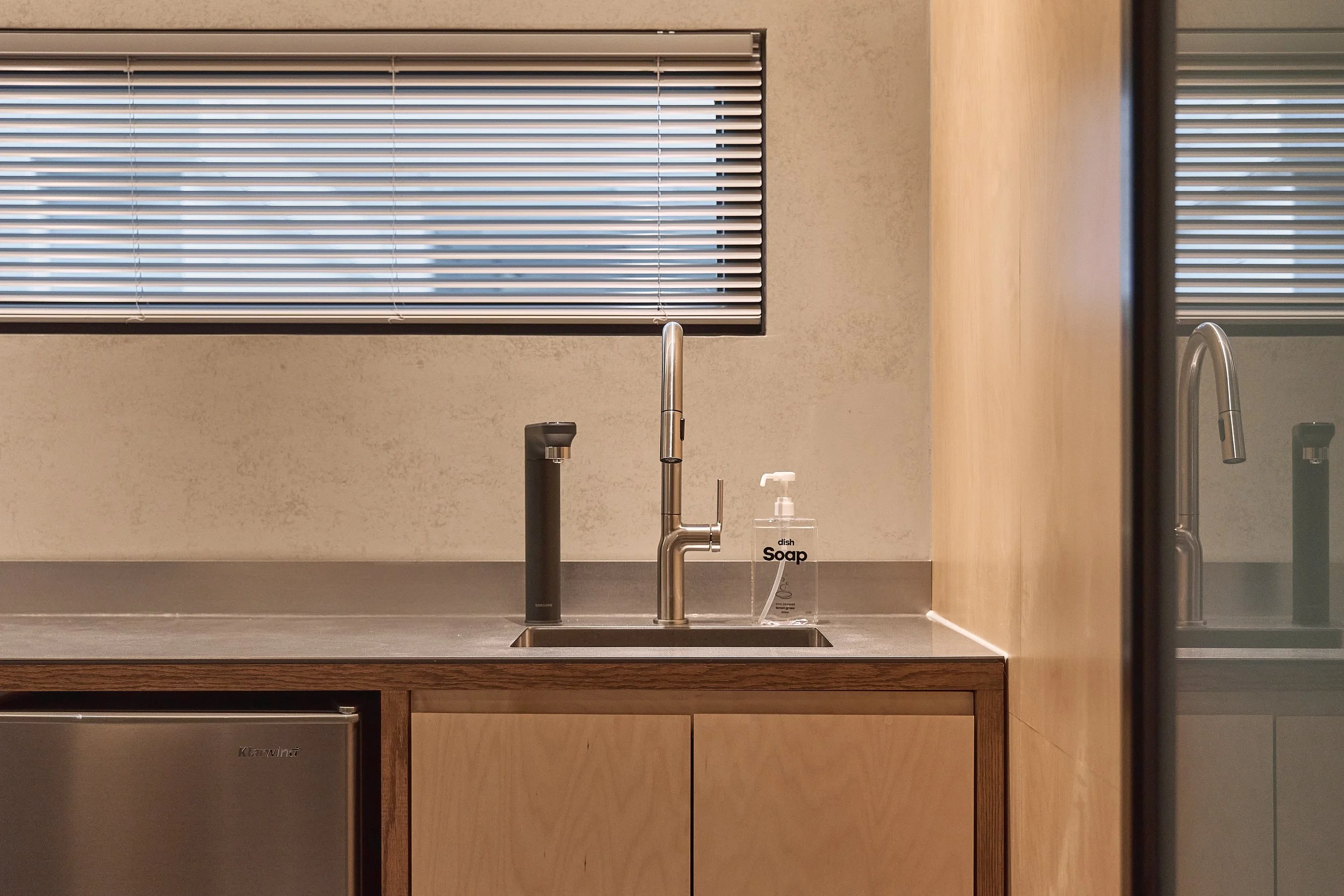 willow leaf Gangnam Room 201 kitchenette with stainless-steel sink, modern faucet, dish soap, and compact Klug mini-fridge in a warm wood and minimalist interior.