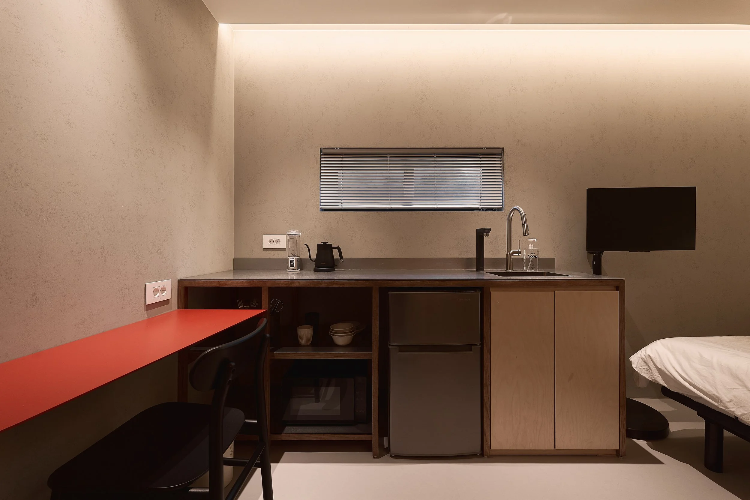 A minimalist willow leaf Gangnam studio featuring a compact kitchenette, stainless-steel sink, mini refrigerator, modern appliances, and a slim red work desk beside the bed.