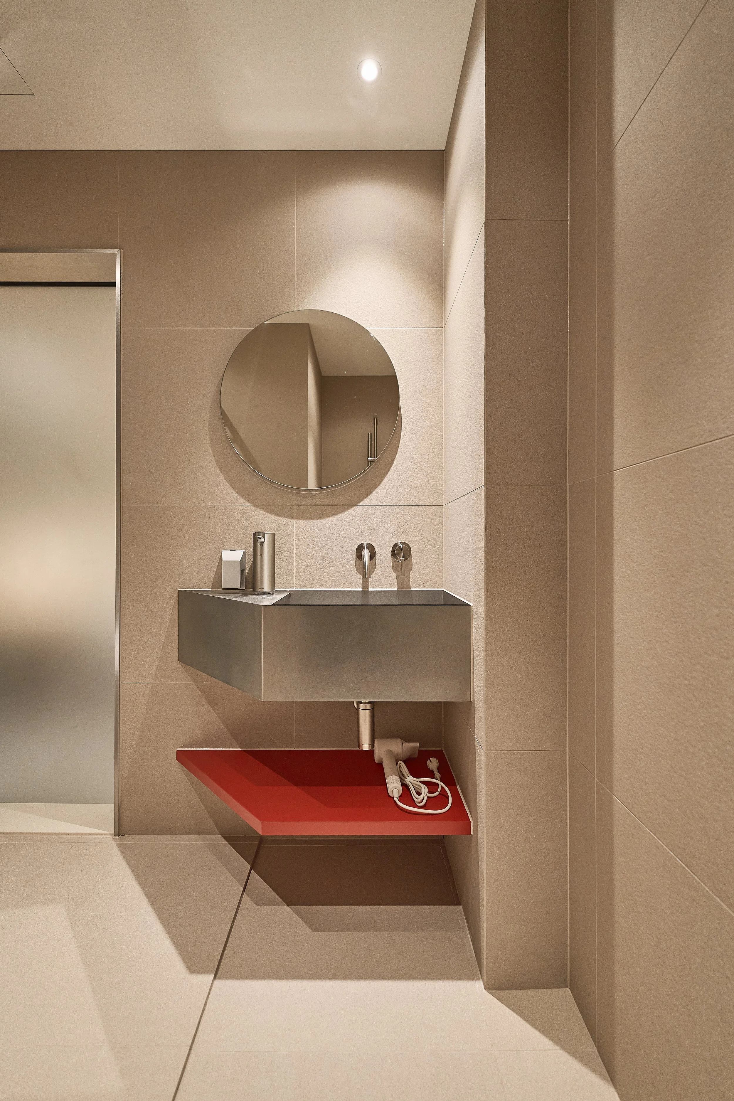 Modern bathroom vanity in willow leaf Gangnam Room 201, featuring a minimalist stainless-steel sink, round mirror, warm beige tiles, and a red shelf with a hair dryer.