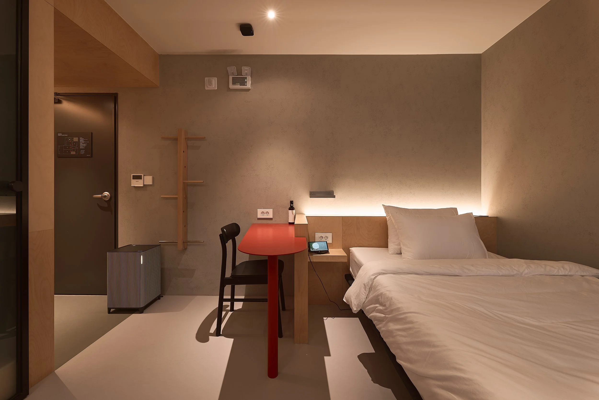 willow leaf Gangnam Room 201 featuring a minimalist bedroom with a cozy bed, warm indirect lighting, red work desk, black chair, wall hooks, and a compact air purifier.