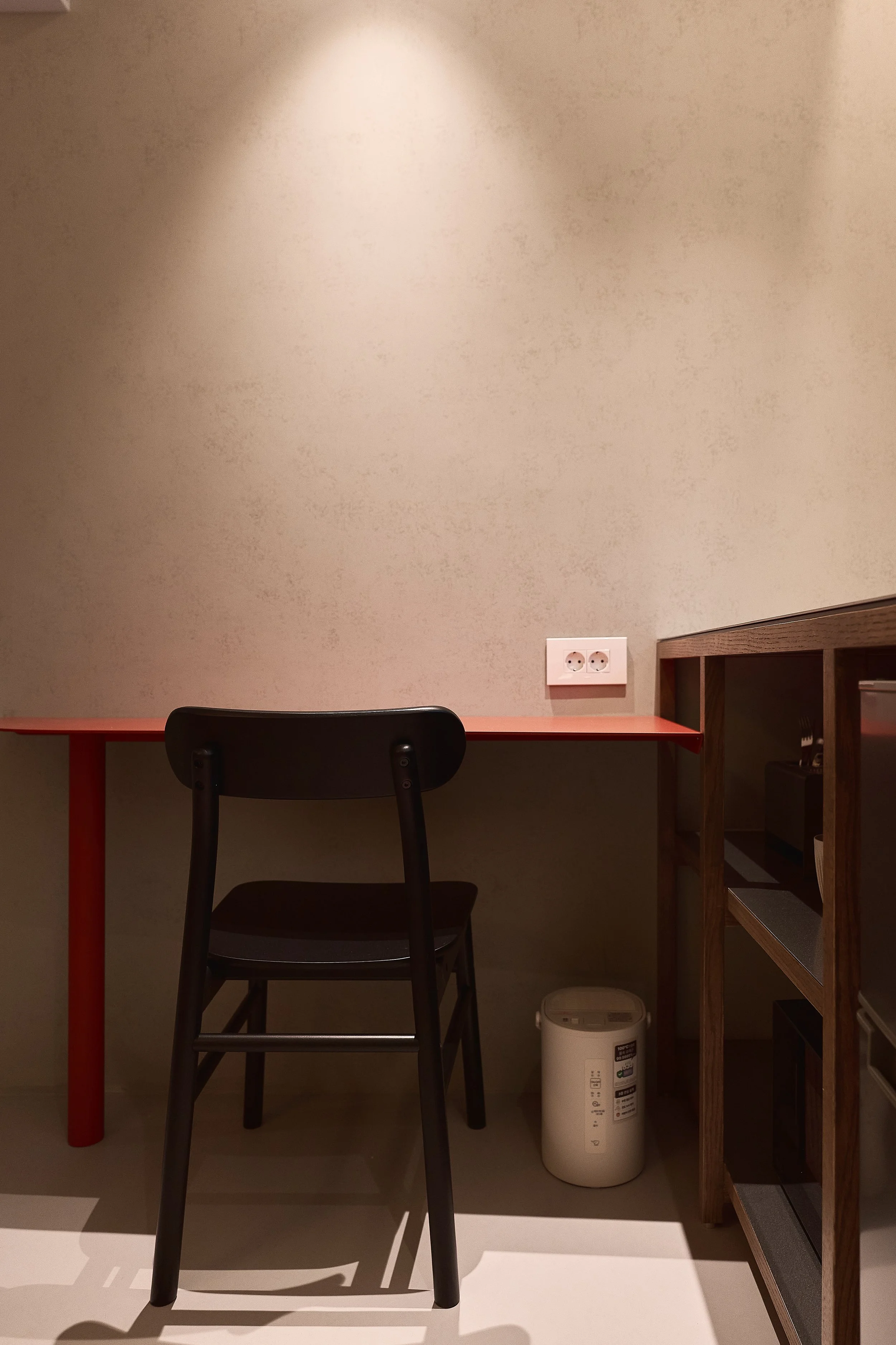 A minimalist work corner in a willow leaf Gangnam room featuring a slim red desk, black chair, soft wall lighting, and neatly organized shelving.