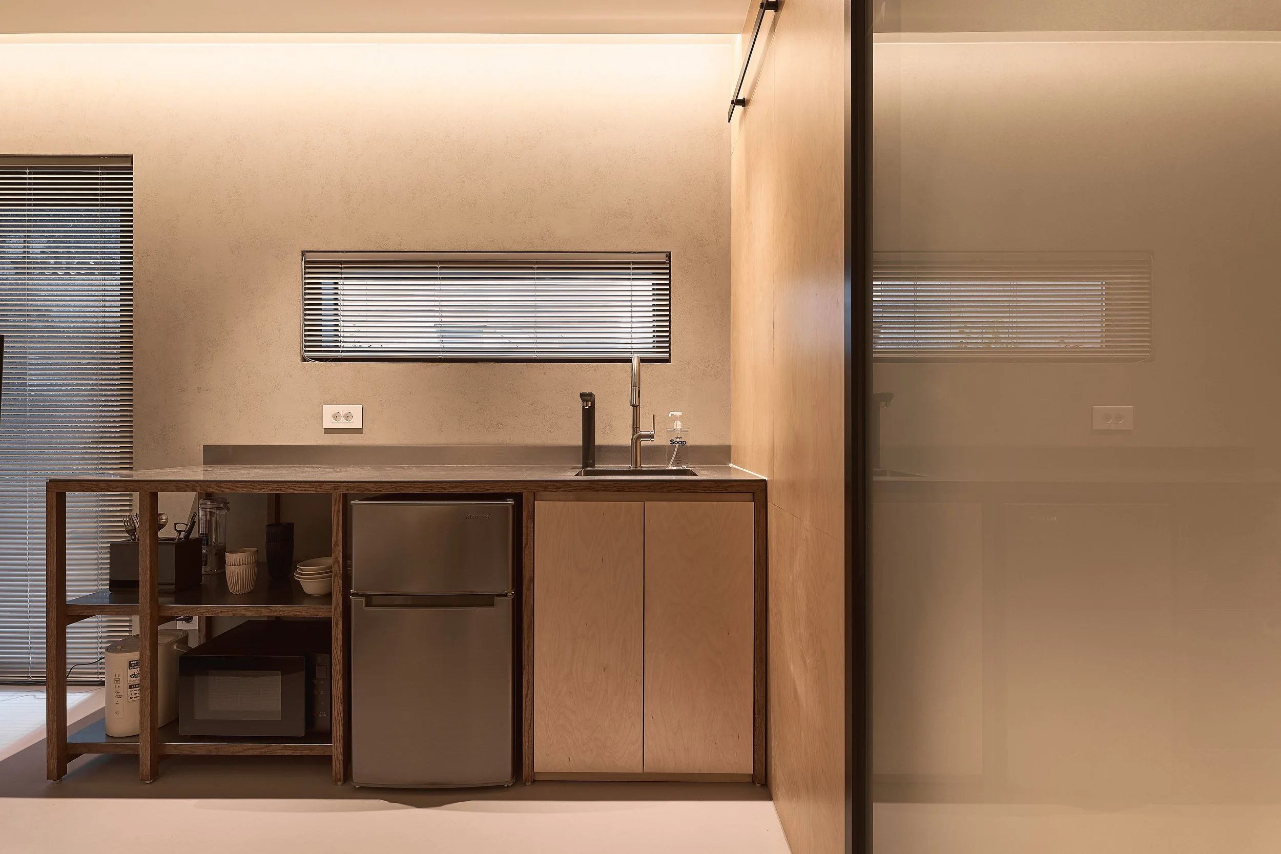 willow leaf Gangnam Room 201 room kitchenette with stainless-steel sink, compact refrigerator, microwave, utensils, and minimalist wood shelving in a warm, modern interior.