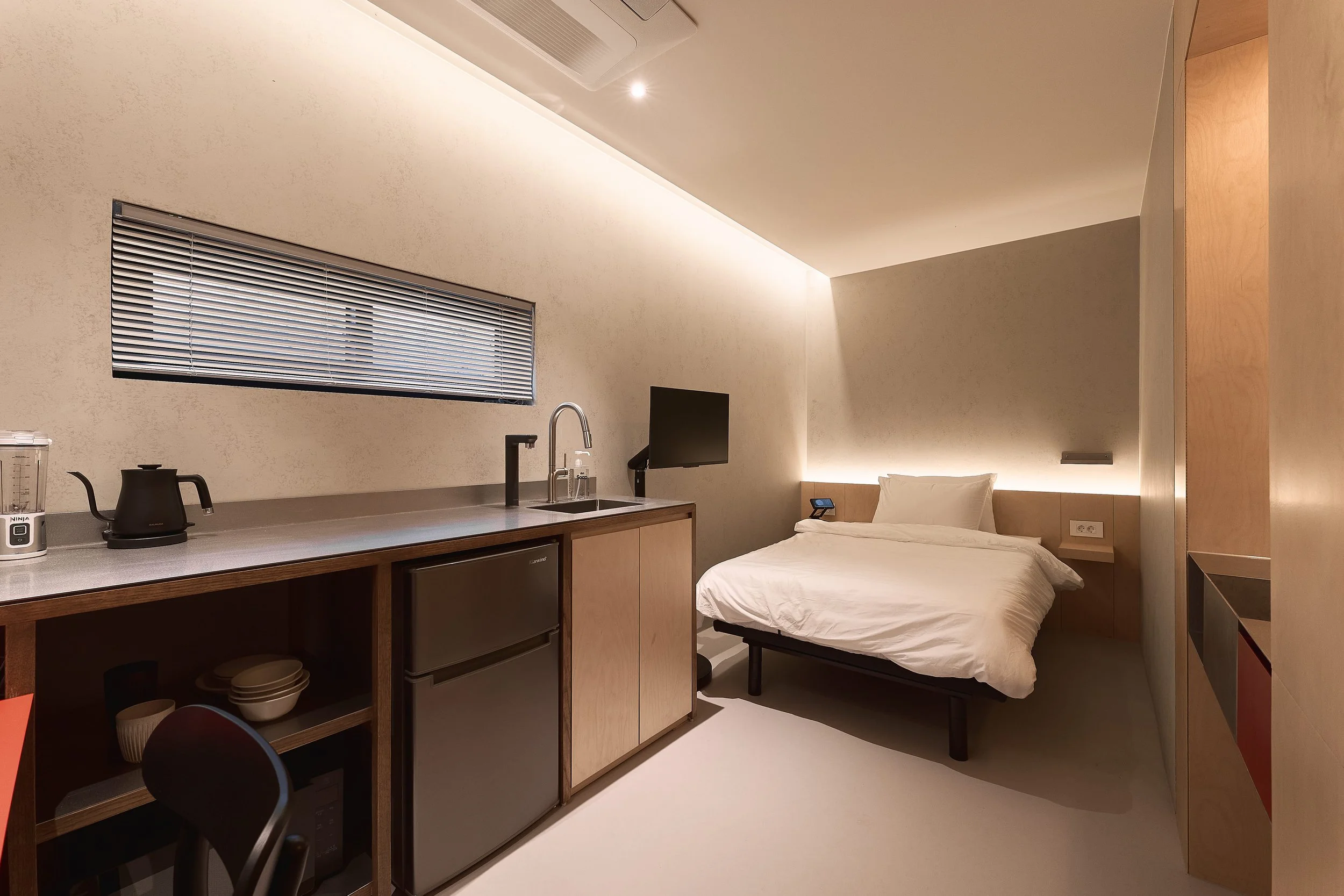A minimalist willow leaf Gangnam studio room featuring a cozy single bed, built-in kitchenette, stainless-steel countertop, compact fridge, and warm indirect lighting.