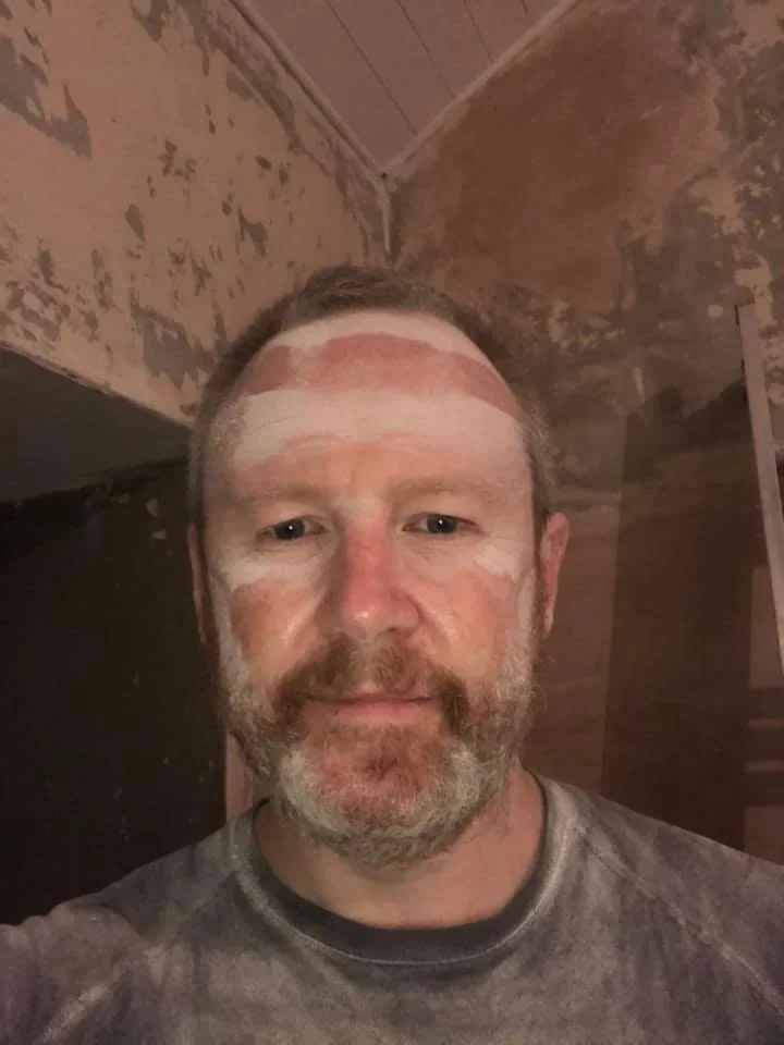 A man with face paint on his forehead, cheeks, and chin, standing in a room with peeling paint on the walls.