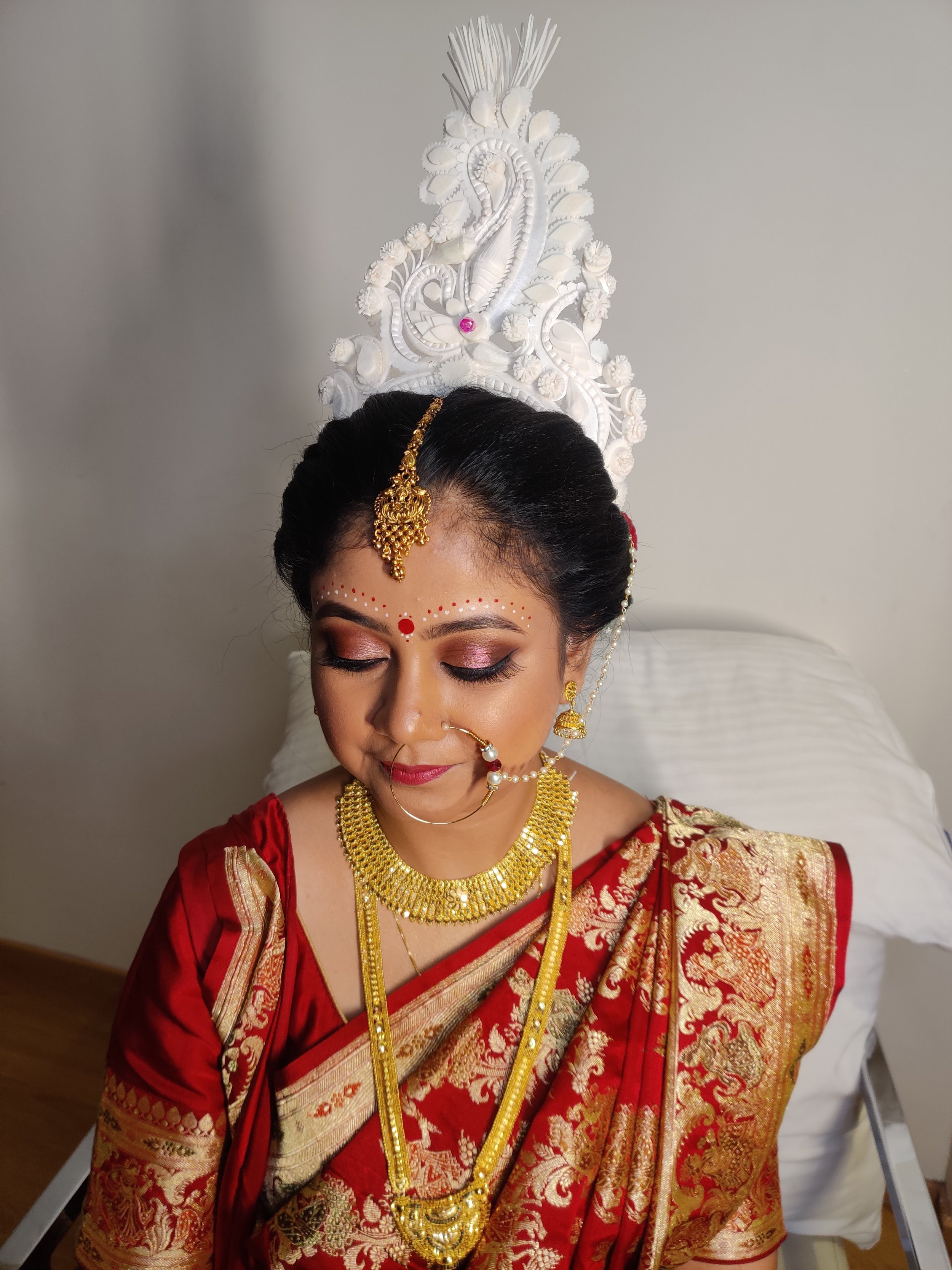 A woman dressed in traditional Indian bridal attire, wearing a red saree with gold embroidery, adorned with gold jewelry including necklaces, earrings, nose ring, and headpiece, with her eyes closed and head bowed, sitting on a bed with a white decor