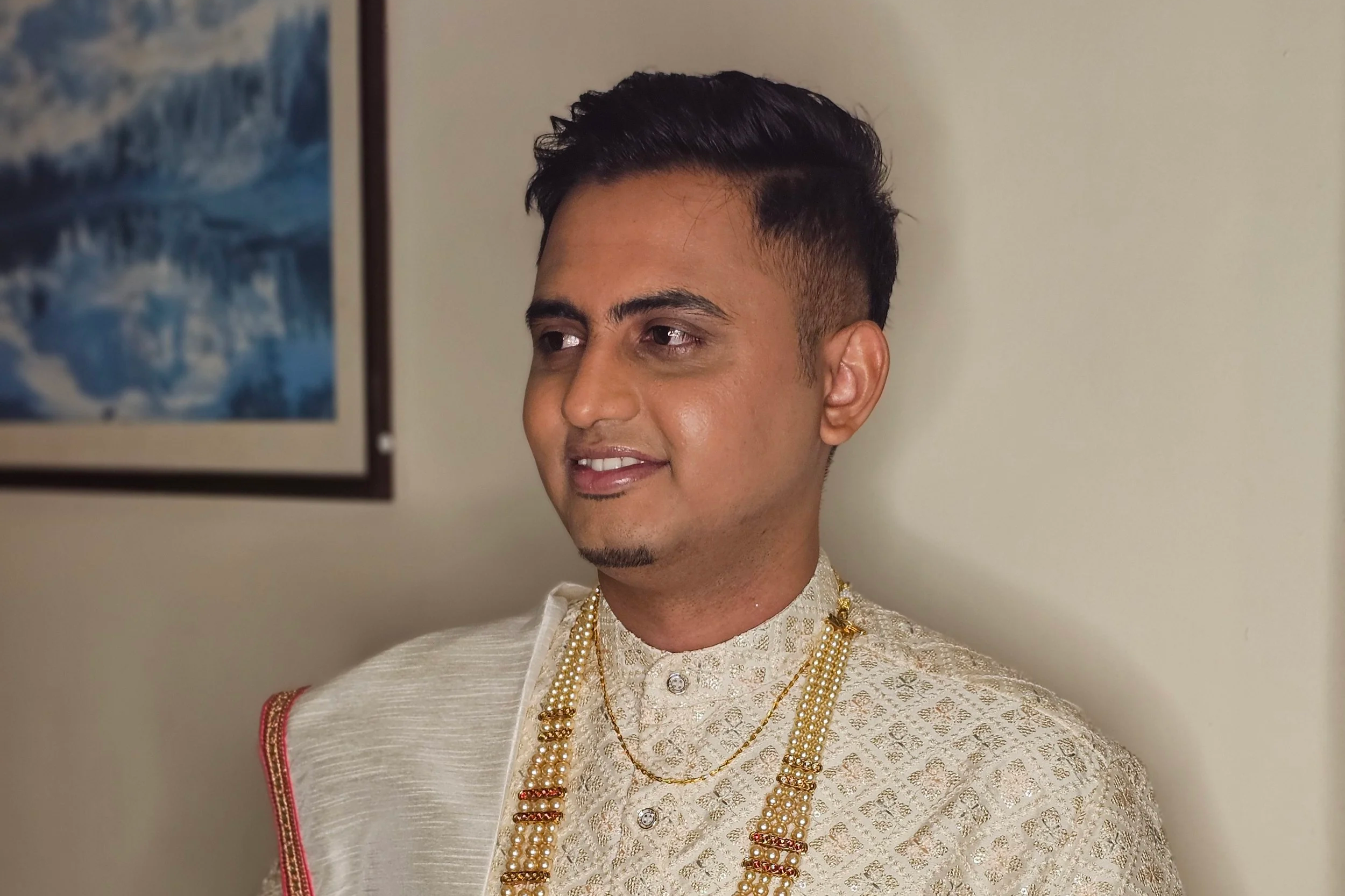 A man dressed in traditional Indian wedding attire with cream-colored sherwani and multiple pearl and gold necklaces, standing against a plain wall with a framed picture of ocean waves in the background.